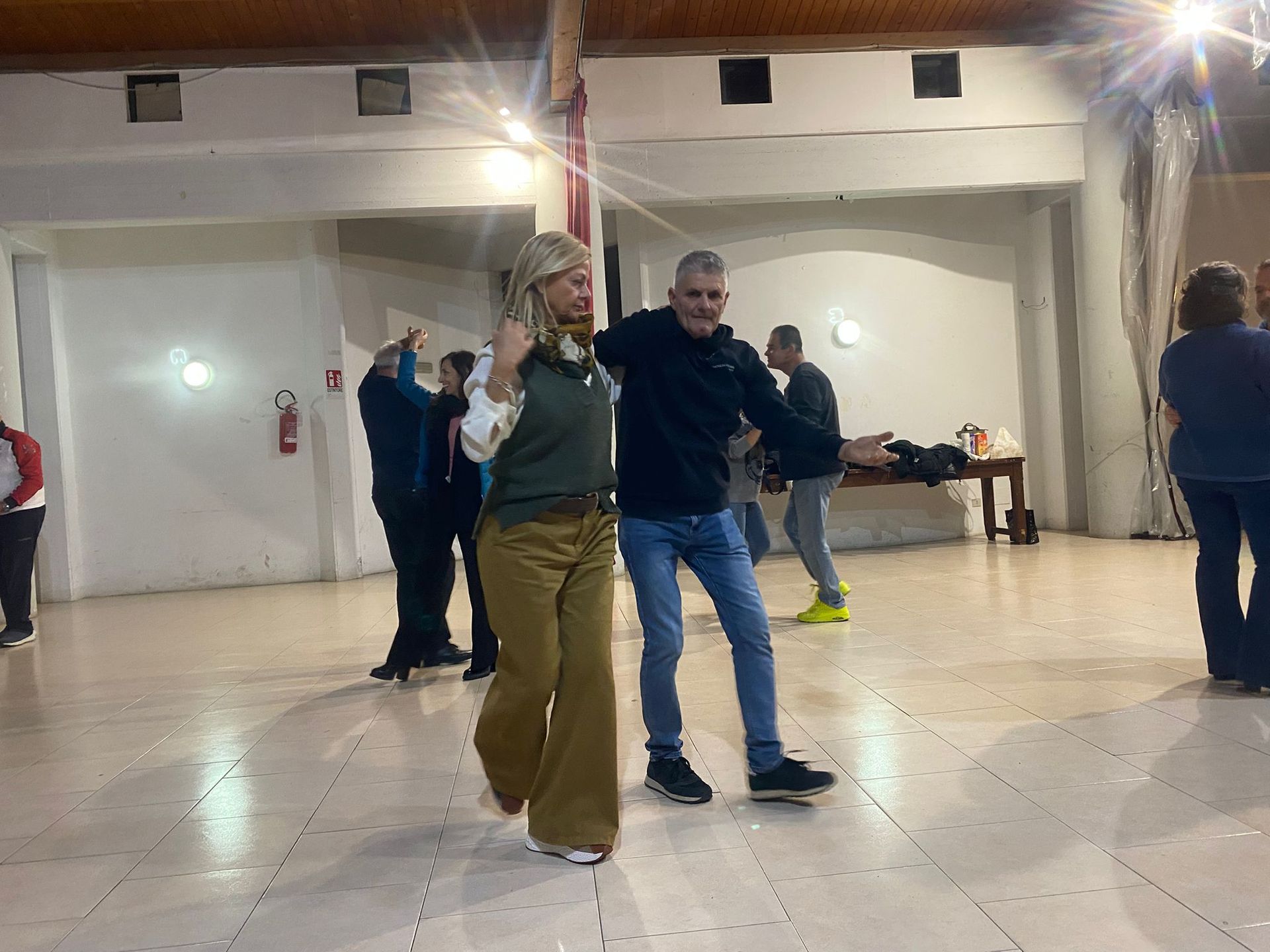 Una coppia balla in una grande sala, sotto gli occhi di altri. Uno indossa una maglietta verde, l'altro una felpa nera con cappuccio.