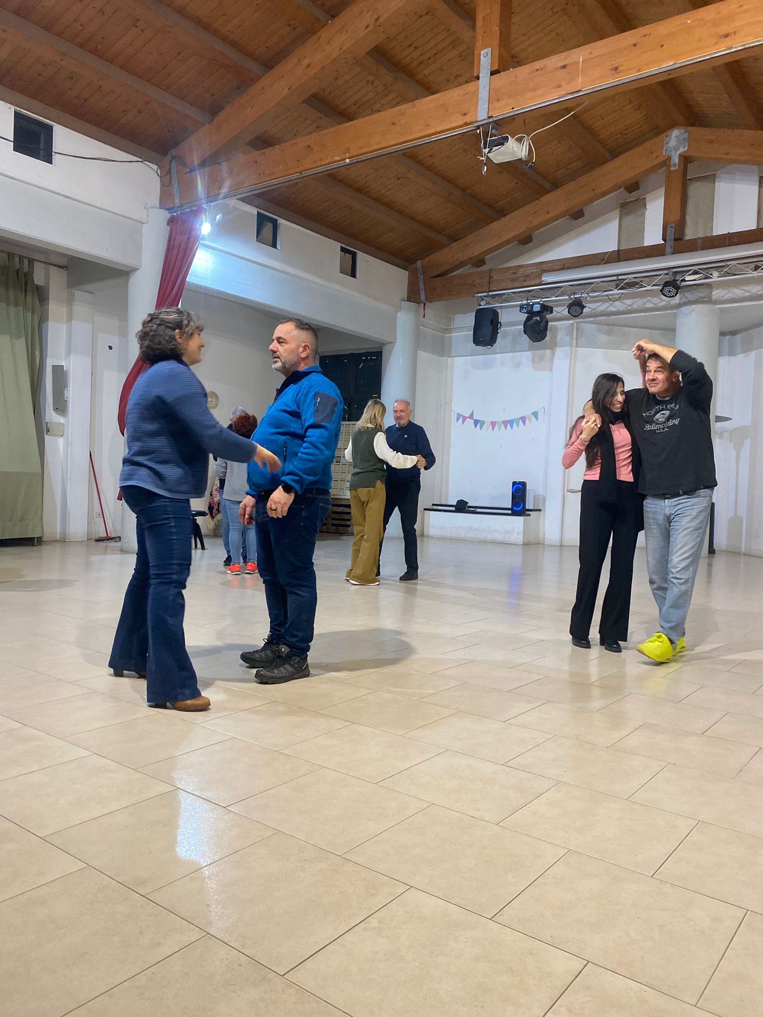 Persone in una sala da ballo, alcune sembrano danzare. Un uomo e una donna interagiscono a stretto contatto al centro della sala.