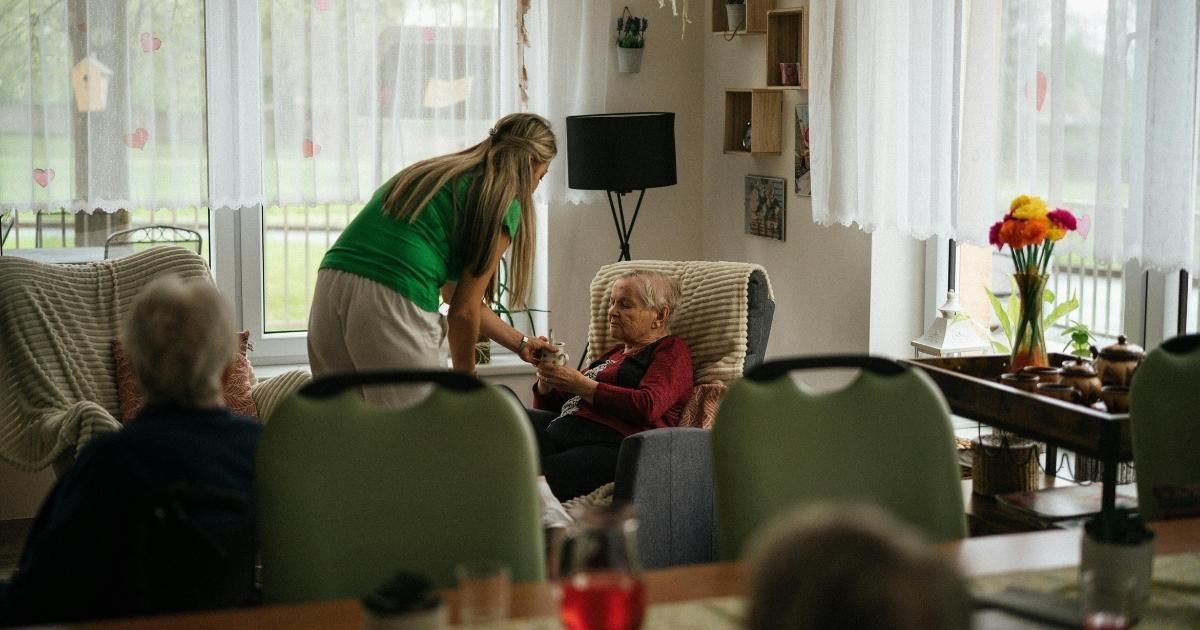 Woman in green top assisting an older person sitting in a chair. Others in the room. Bright room with flowers.