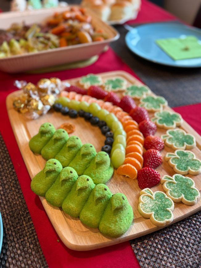 St. Patrick's Day charcuterie: green chick treats, rainbow fruit, clover cookies, and a main course in the background.