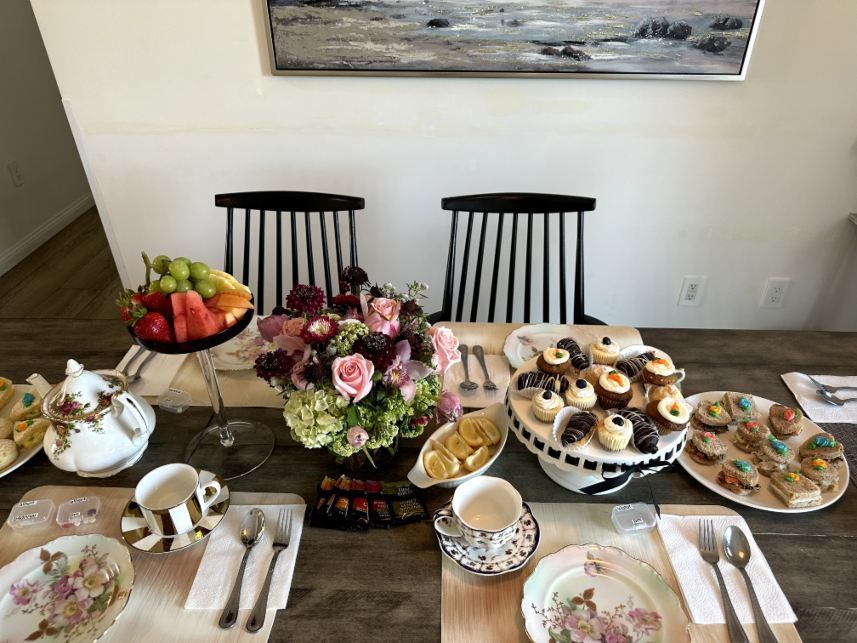 Tea party set for two: flowers, desserts, fruit, ornate china on a wooden table, striped chairs in the background.