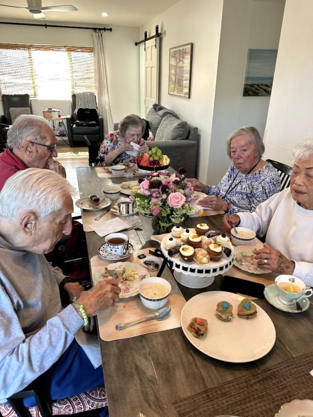 Elderly group at a table, enjoying tea and pastries. Sunlit room, focused expressions, vibrant flowers, treats.