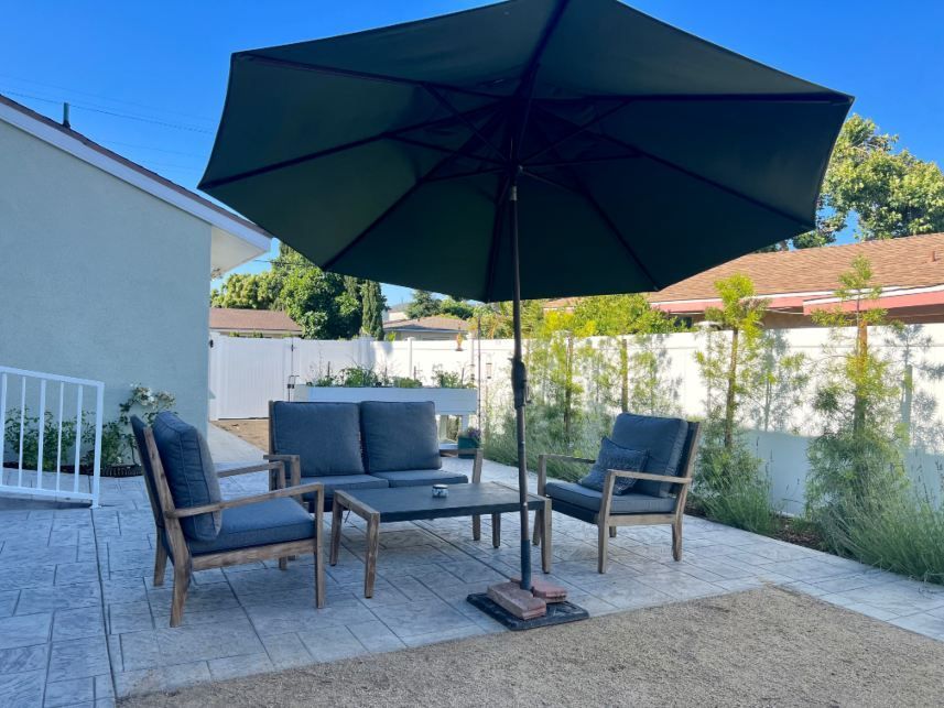 Patio set with umbrella, two chairs, and a sofa on a concrete patio, surrounded by greenery.