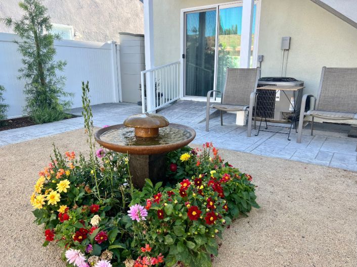 Fountain surrounded by colorful flowers in a gravel garden, with patio and house in the background.