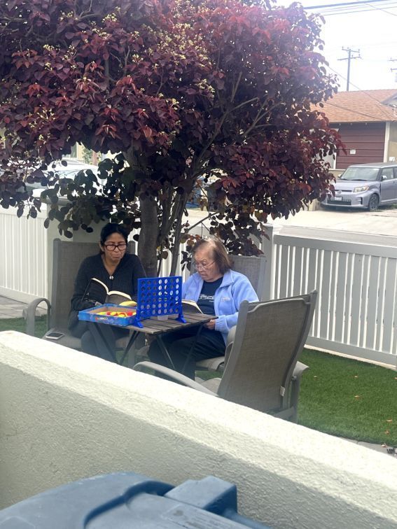 Two women sit outdoors under a tree, playing a board game and reading. Coral Tree Care Home in Lomita, CA.