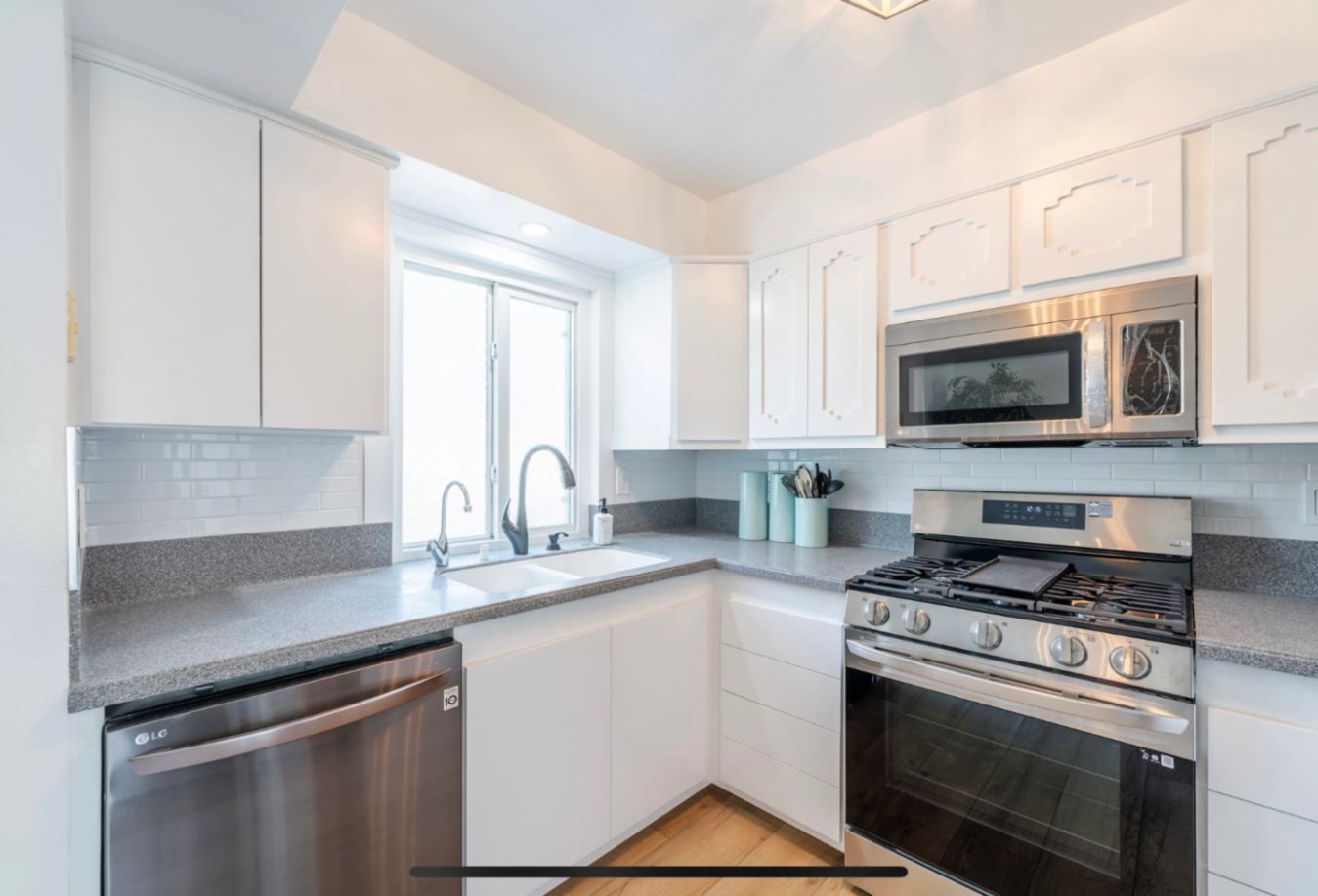 White kitchen with stainless steel appliances, gray countertops, and a window.