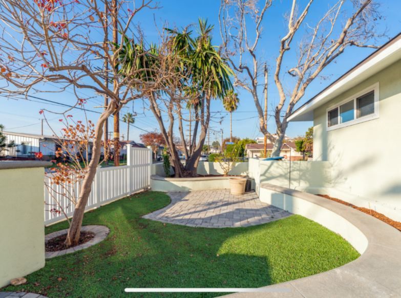 Lush green lawn and curved pathway lead to a brick patio, trees and a light green house with blue sky.