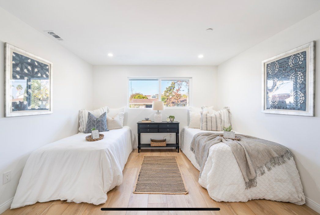 Twin beds in a bright white bedroom with art, a black nightstand, and a rug.