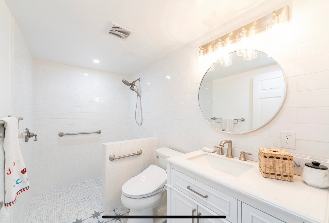 White bathroom with shower, toilet, sink, and round mirror. Includes grab bars for accessibility.