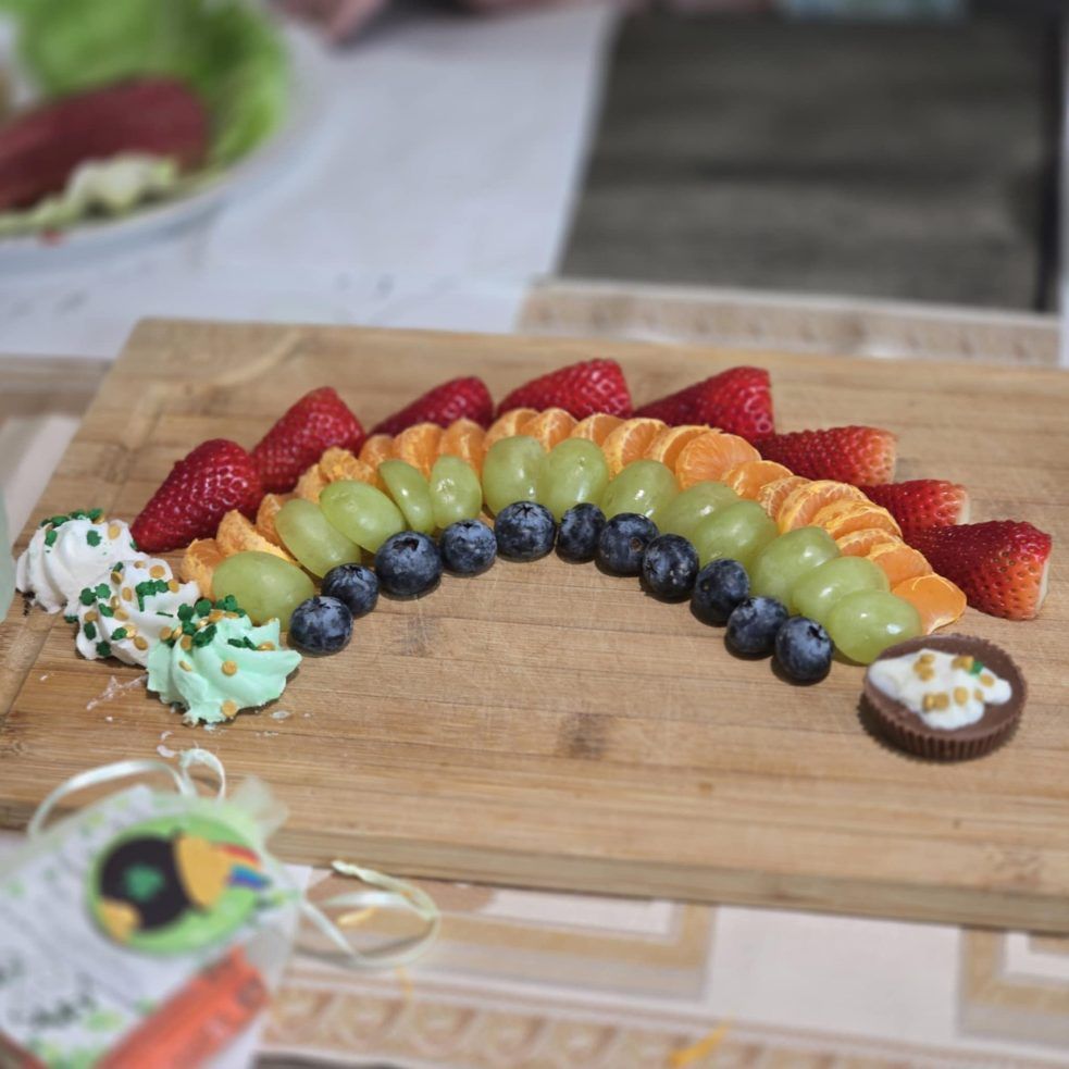 Rainbow fruit platter on a wooden board: strawberries, oranges, grapes, blueberries, and desserts.