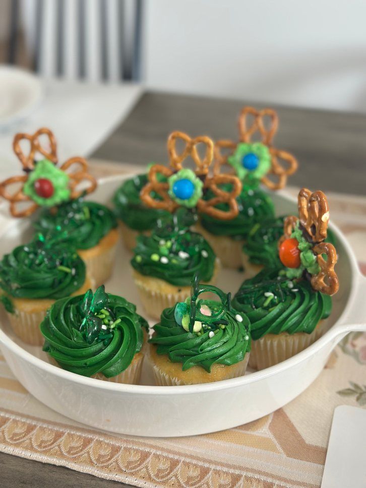Cupcakes with green frosting and pretzel and candy decorations in a white dish on a table.