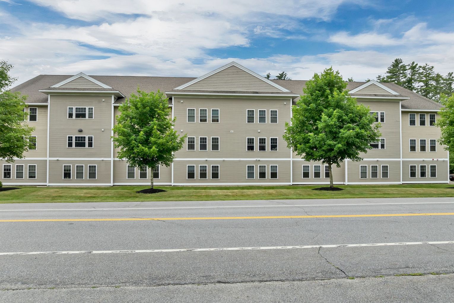 Three-story beige apartment building with trees in front and a road in the foreground