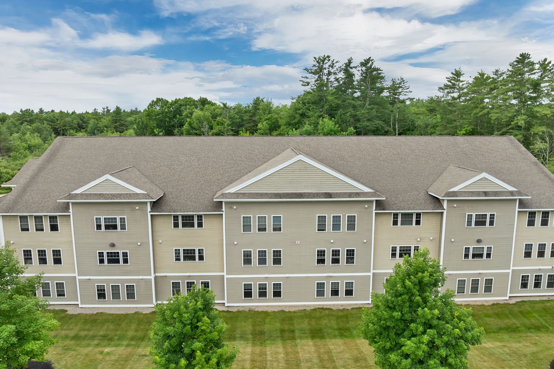 Three-story beige apartment building with white trim, set in a grassy area with trees and a blue sky