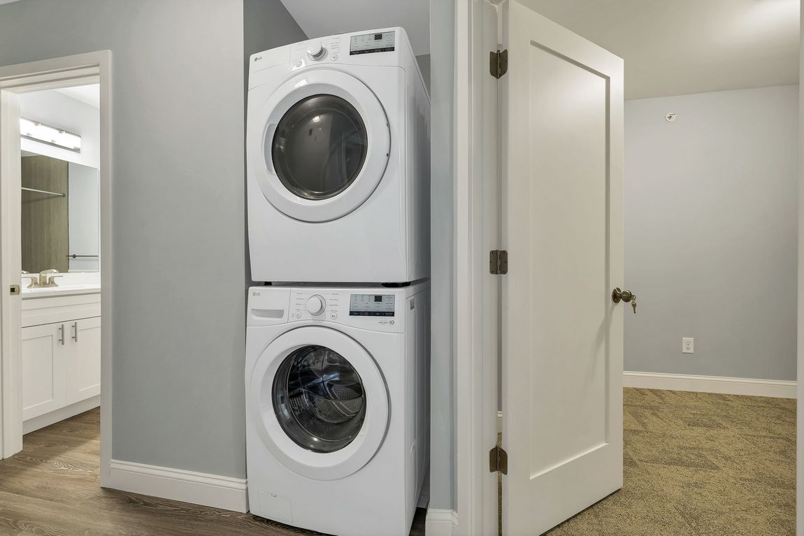 Stacked white washer and dryer in a laundry closet beside an open door, with beige carpet and gray walls.