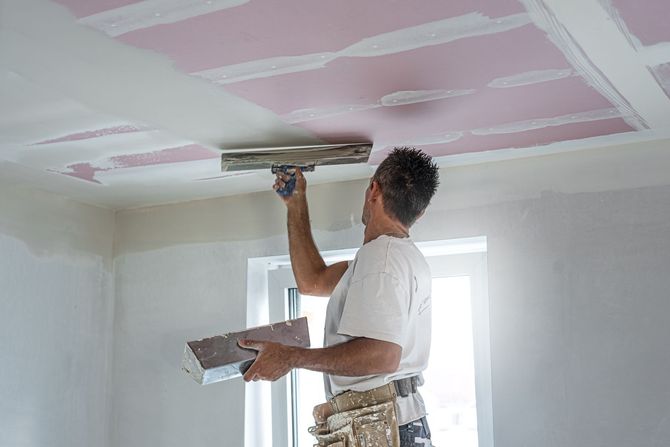Construction worker installing drywall on a ceiling while standing on a scaffold in a room under renovation.
