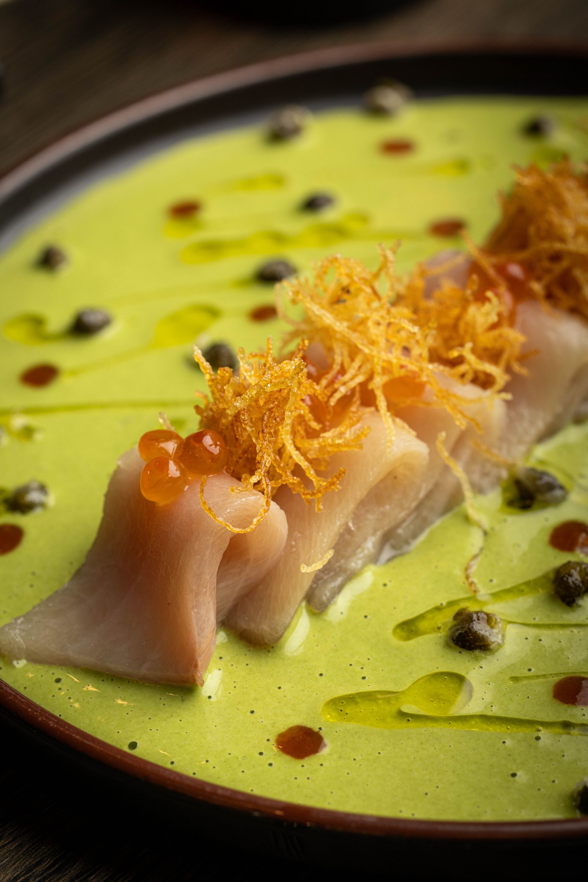 Thin sliced fish on green sauce, topped with crispy garnish and droplets of sauce