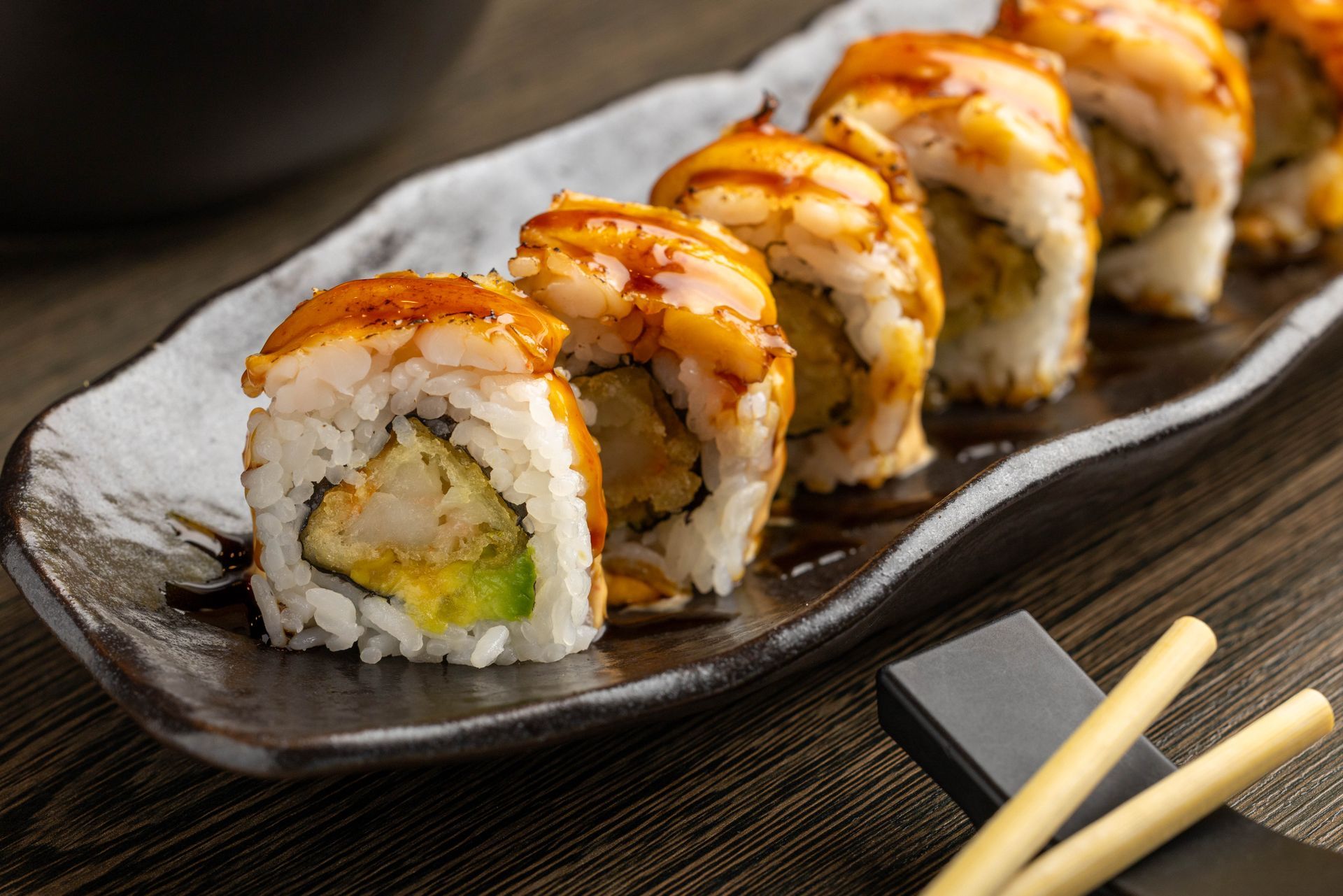 Sushi rolls topped with sauce on a black plate, with chopsticks on a wooden table.