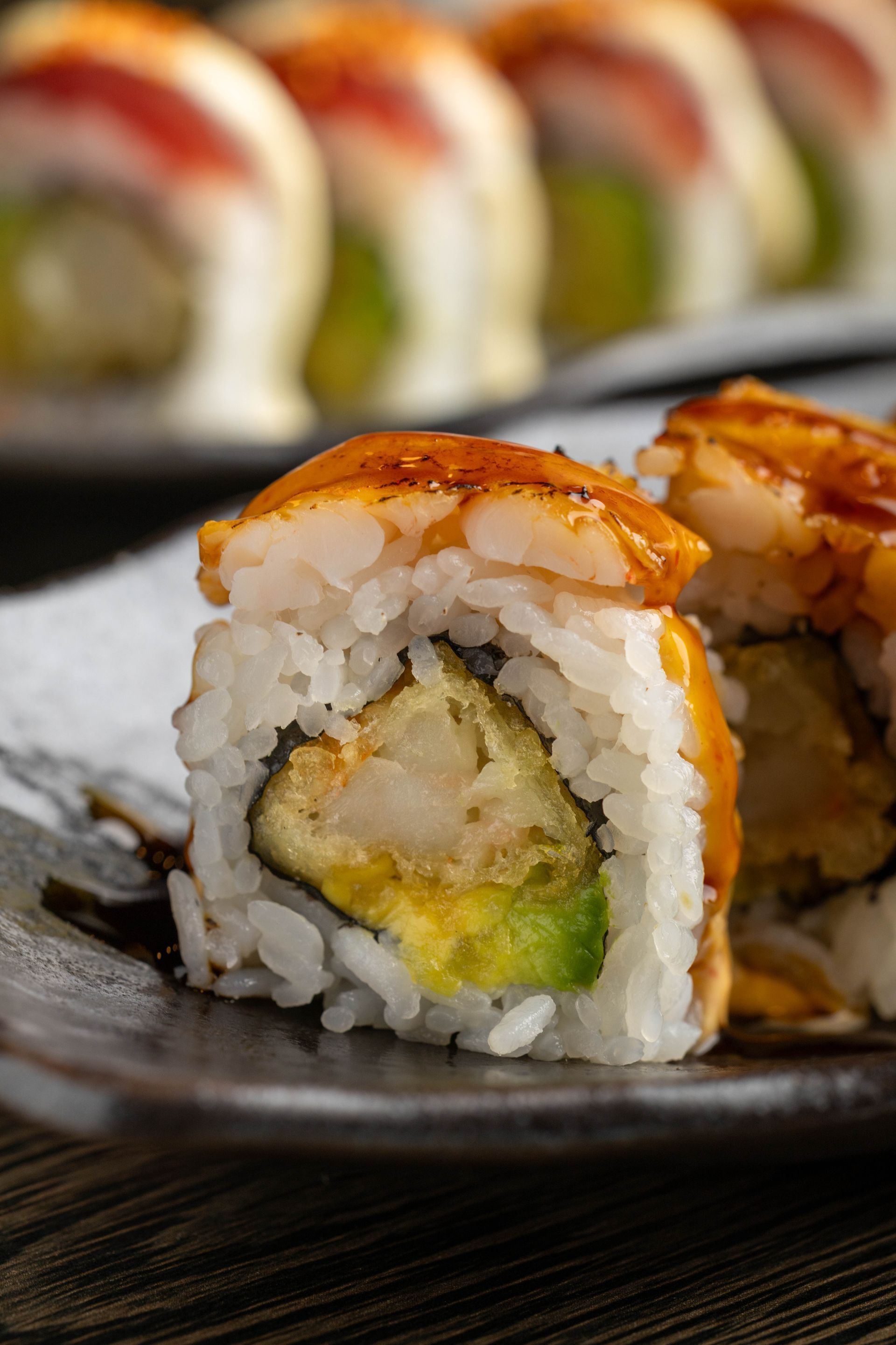 Close-up of sushi rolls with avocado and orange topping on a black plate, with more rolls blurred in the background