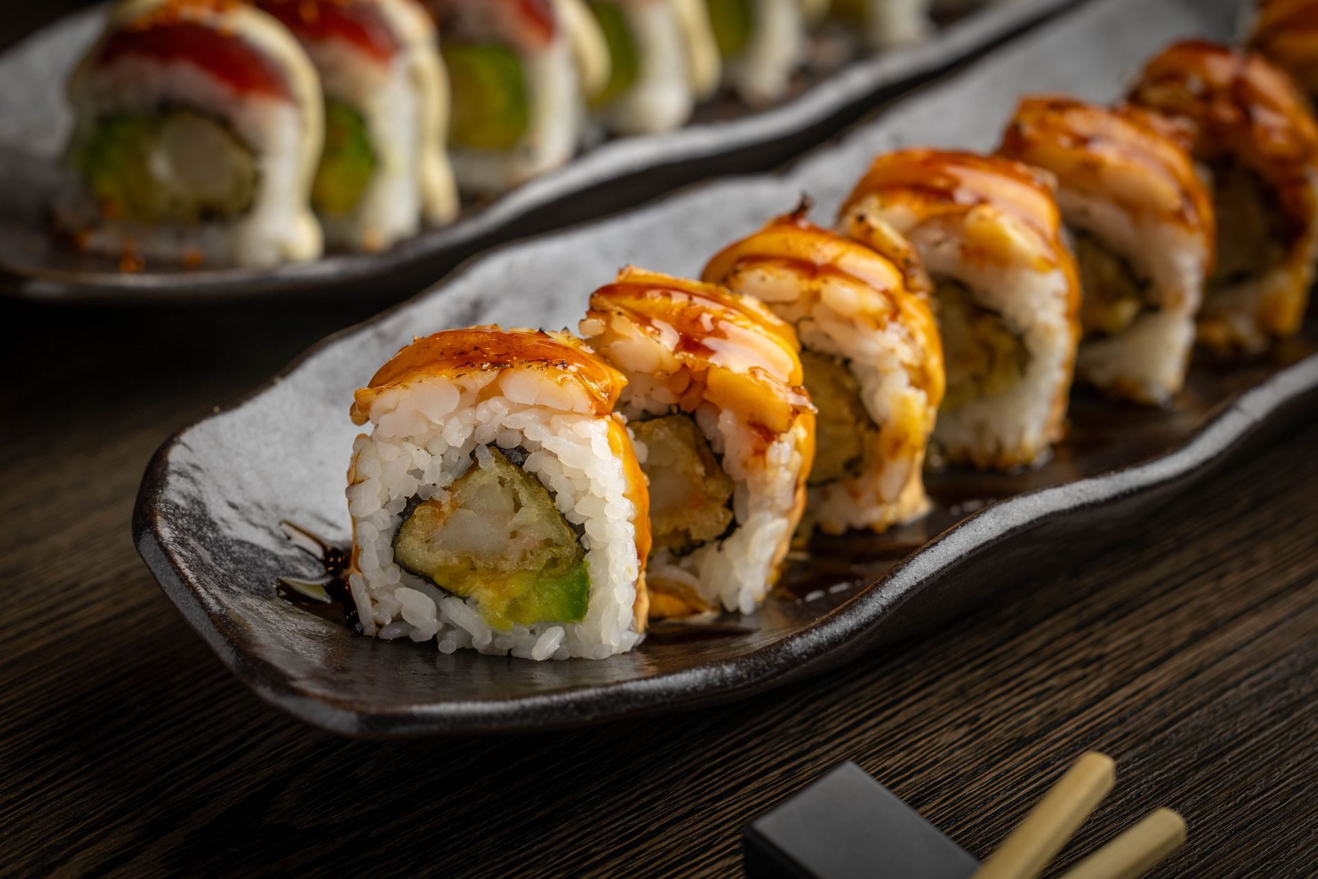 Sushi rolls drizzled with sauce on a dark ceramic plate, with chopsticks nearby
