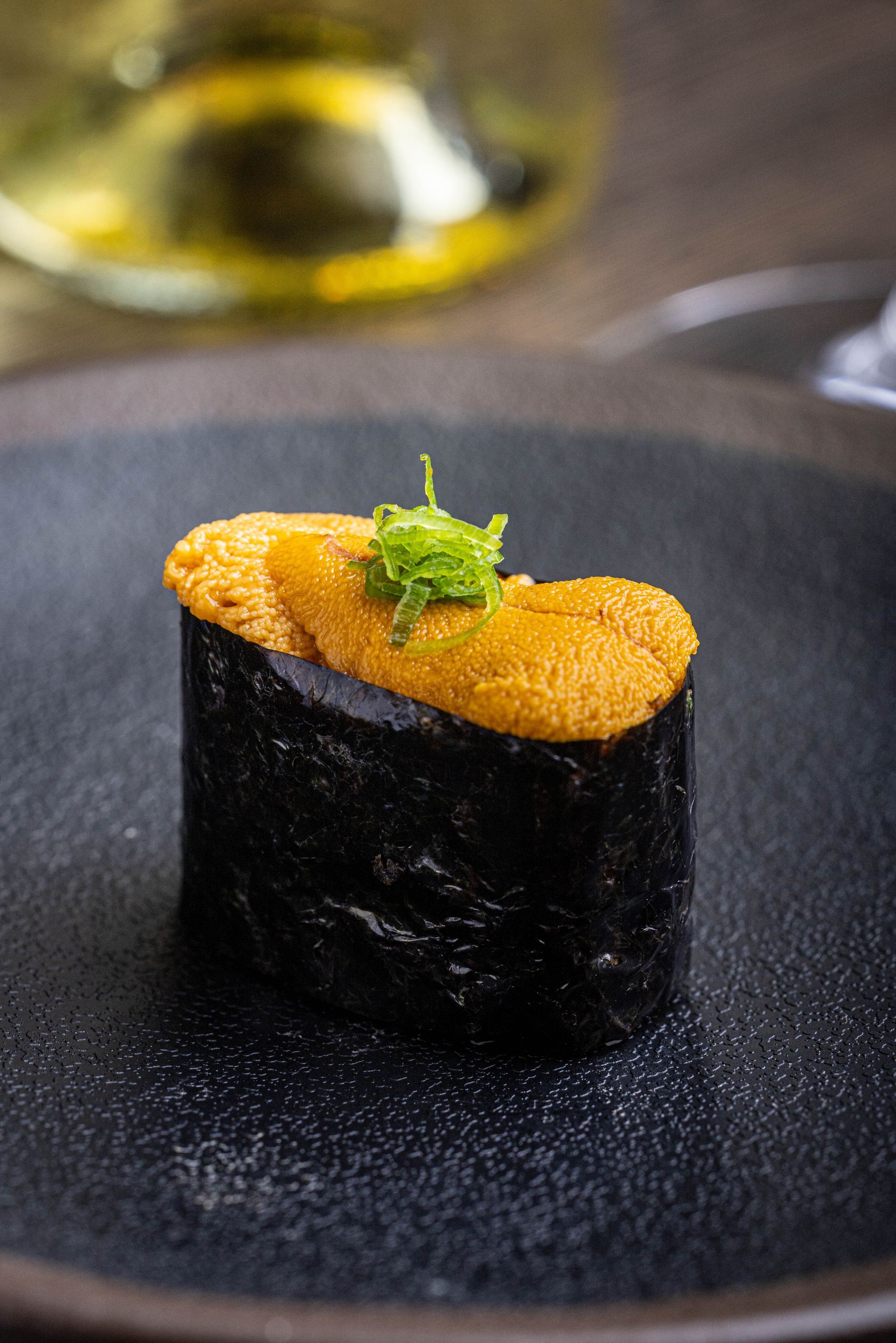 Black nori-wrapped sushi topped with orange roe and green garnish on a dark plate