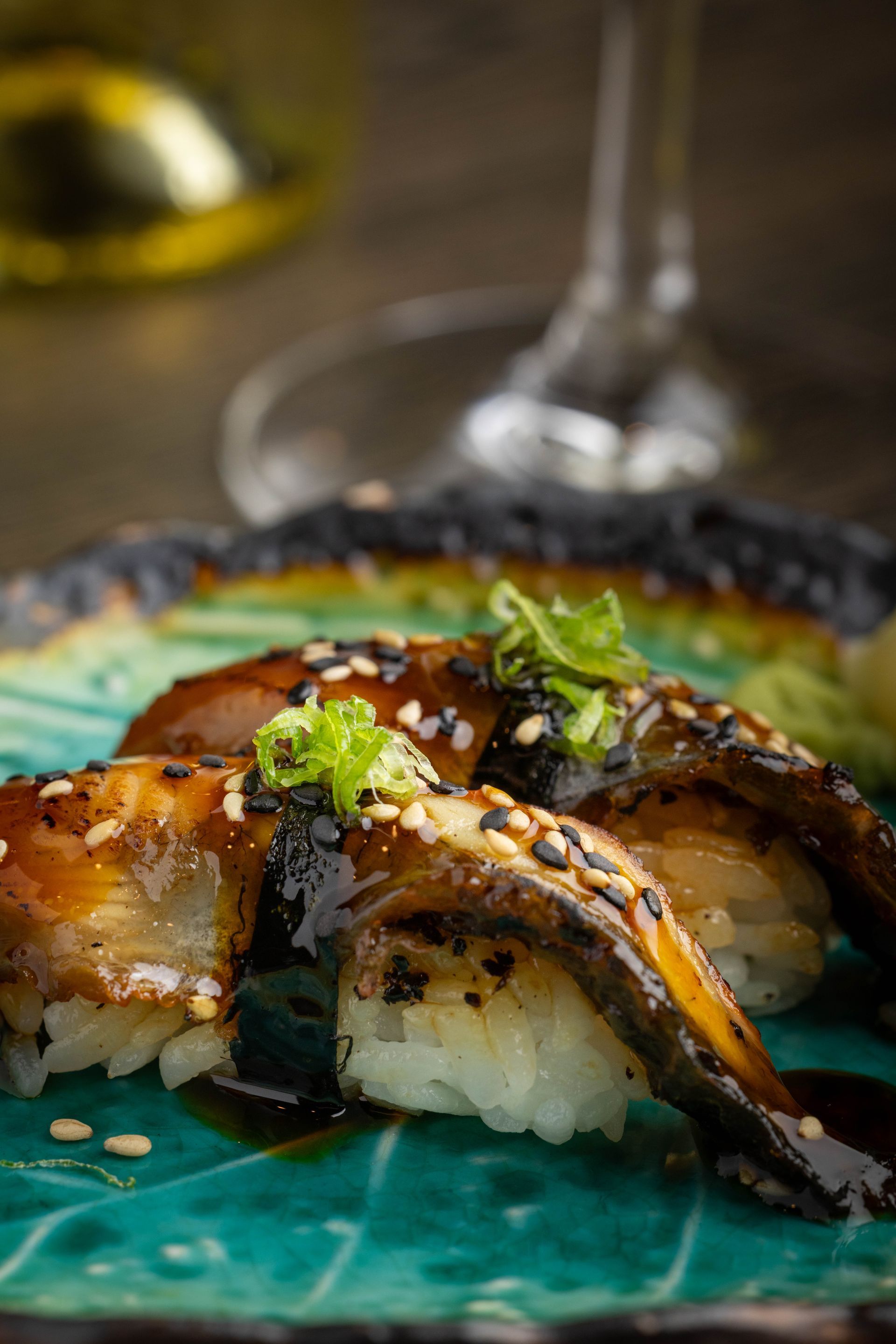 Sushi rolls topped with glazed eel and sesame on a turquoise plate, with chopsticks and a blurred glass in the background