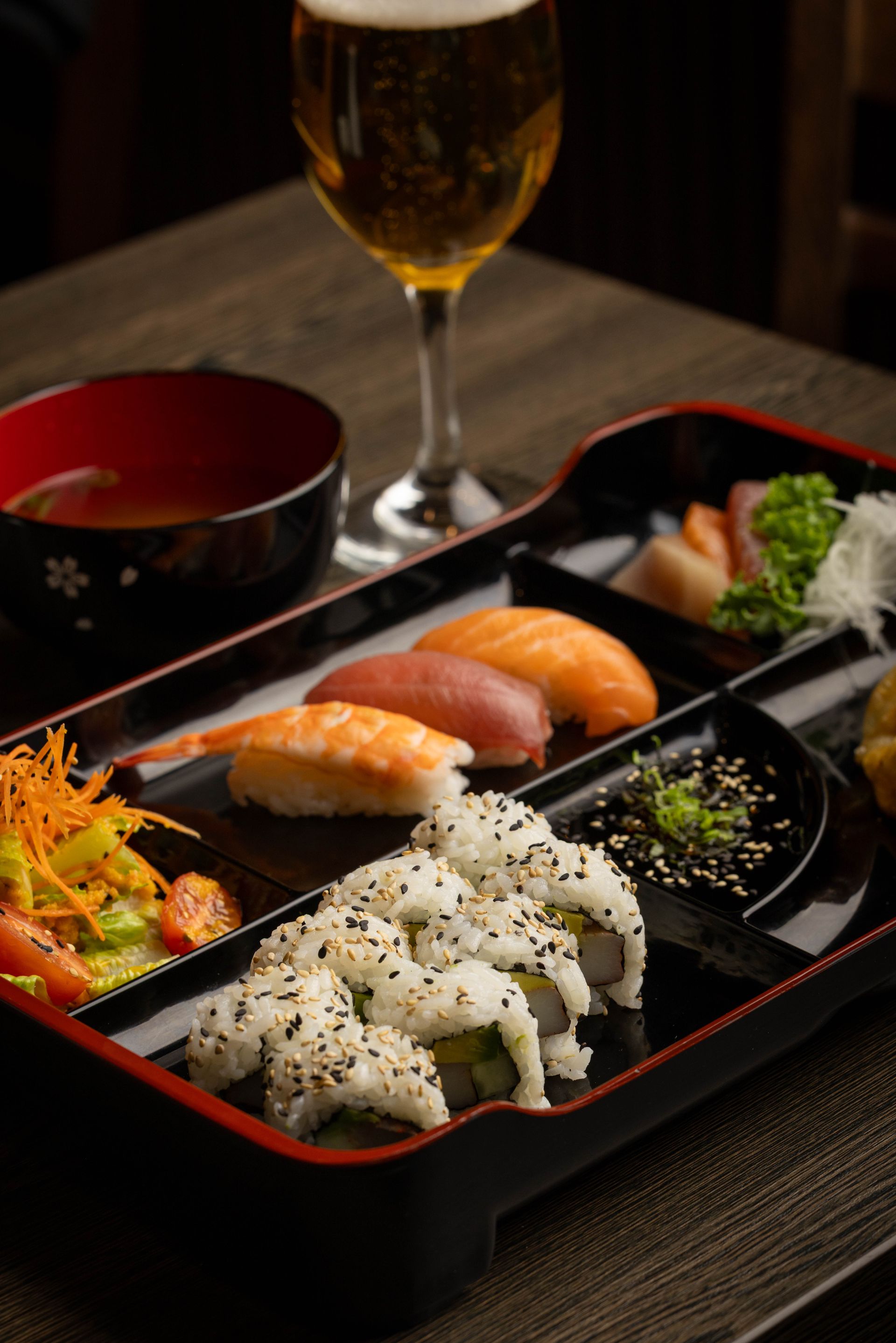 Sushi bento box with nigiri and rolls beside a bowl and a glass of beer on a table