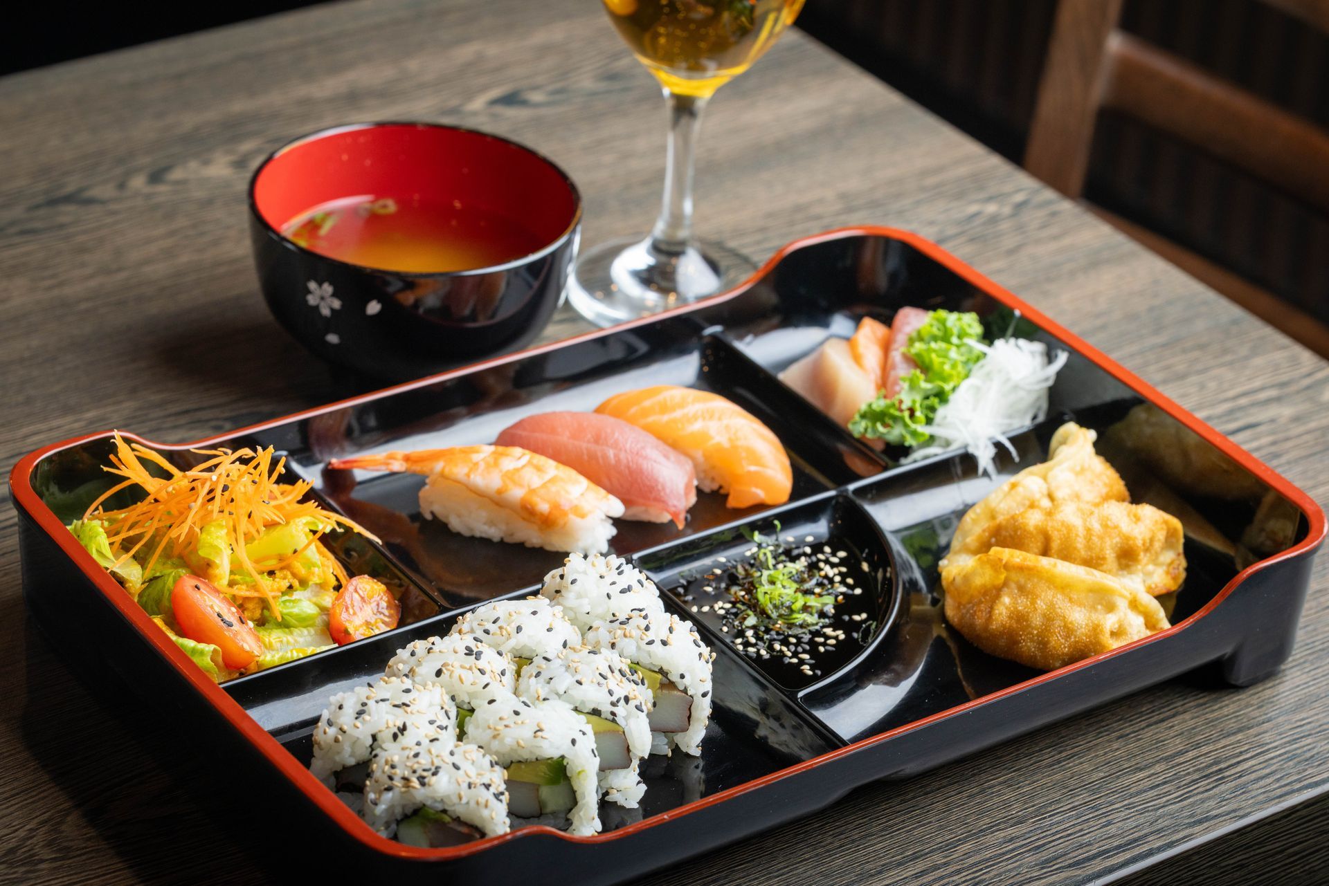 Assorted sushi and Japanese side dishes in a black bento tray with miso soup and a drink
