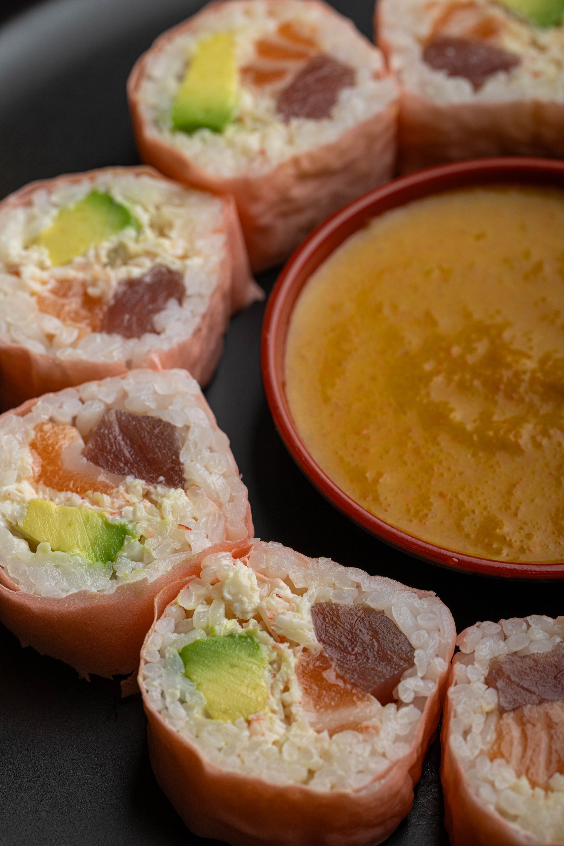 Sushi rolls with avocado and tuna beside a small bowl of dipping sauce on a dark plate
