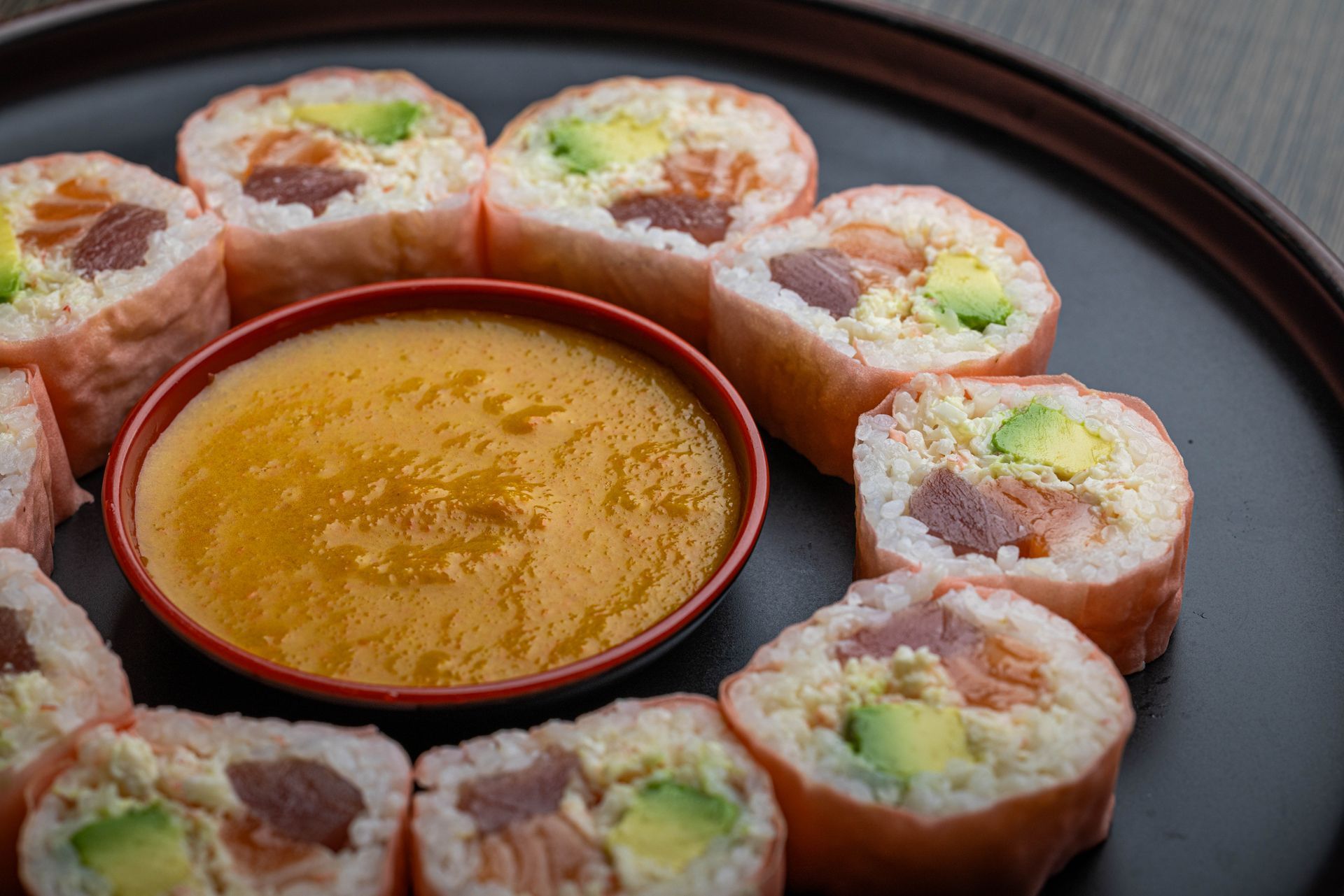 Sushi rolls arranged on a black plate with a small bowl of dipping sauce in the center