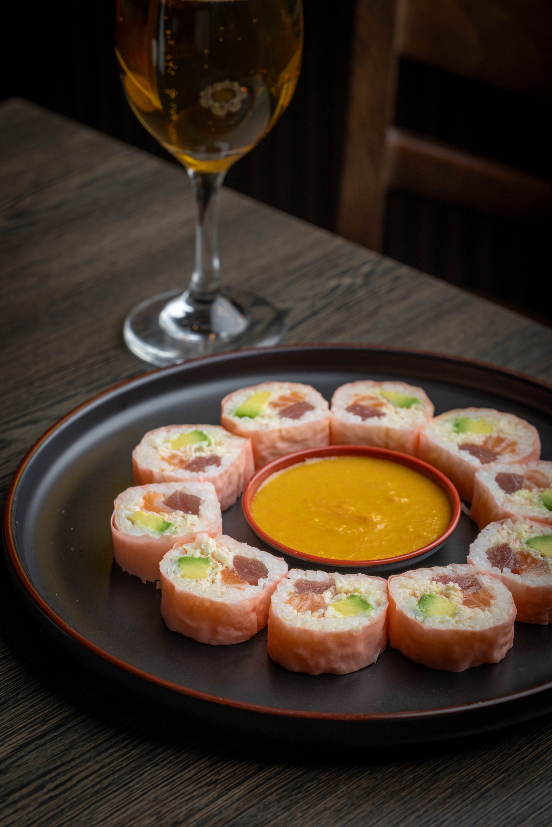 Plated sushi rolls with orange dipping sauce on a dark table beside a wine glass