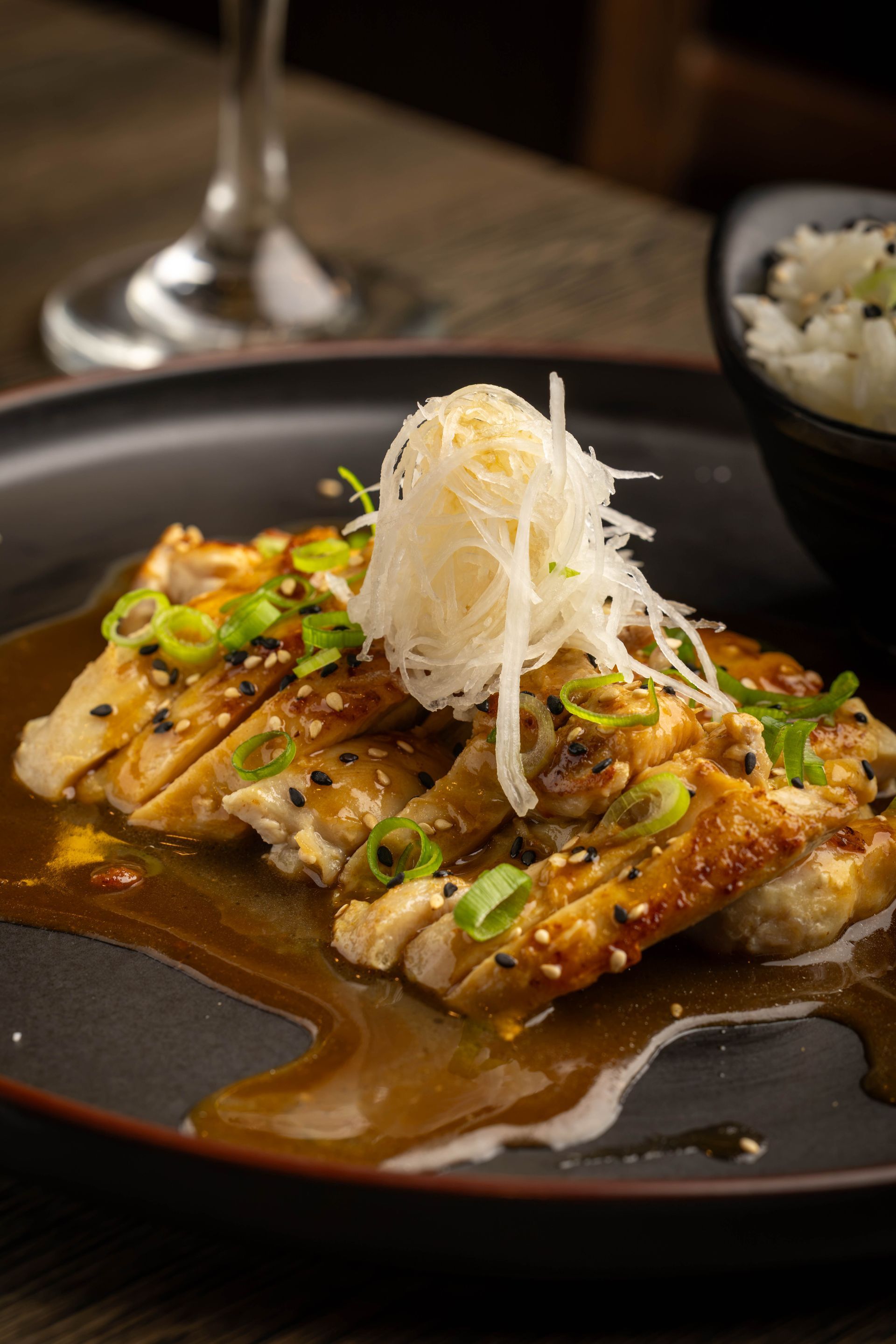 Grilled fish topped with white garnish and green onions on a dark plate, with sauce and rice in the background