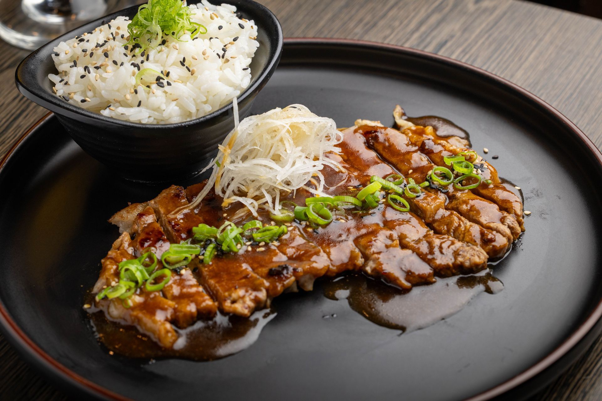 Glazed grilled steak on a black plate with rice and shredded garnish on a dark table