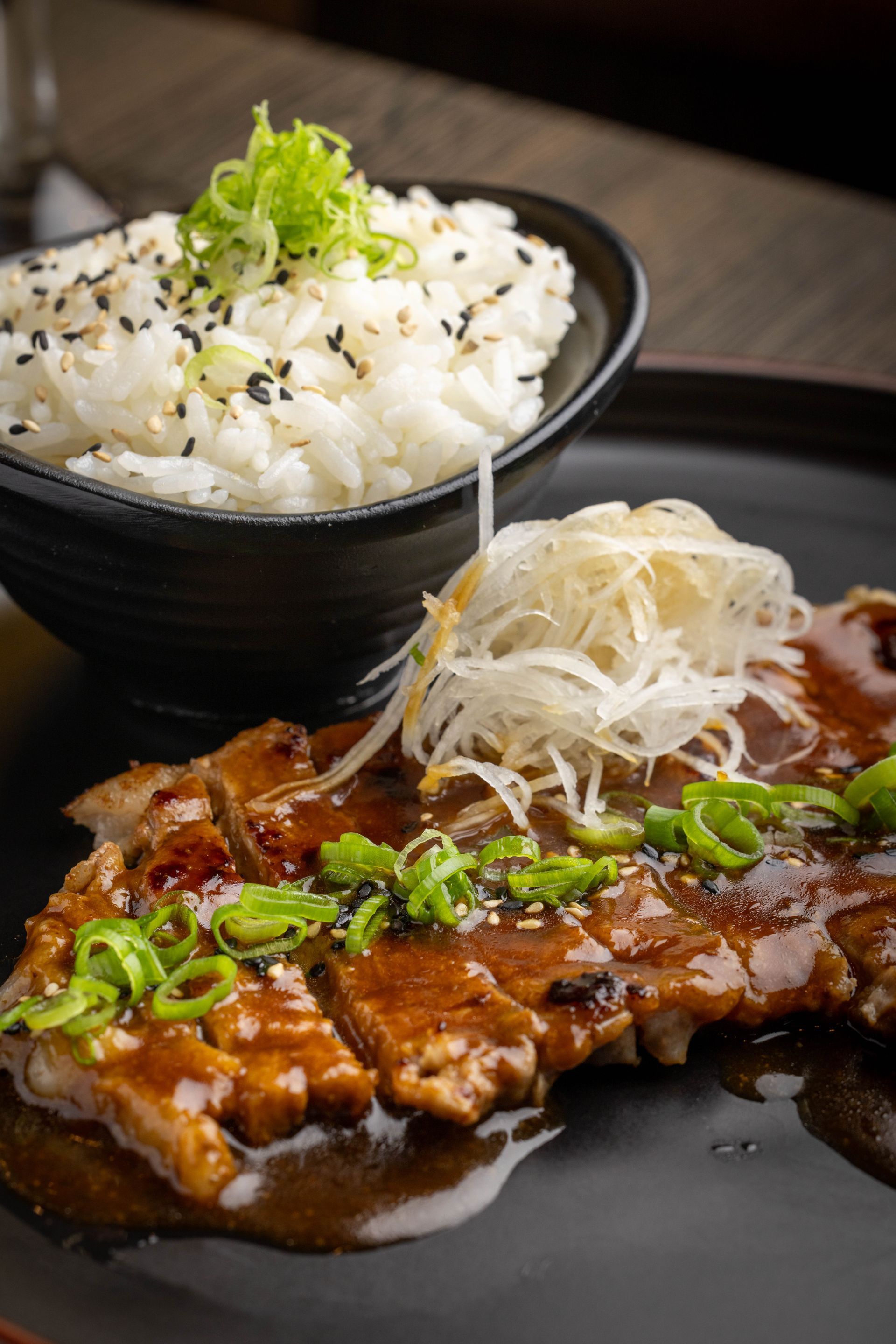 Rice bowl with chopped green garnish and glazed meat topped with sliced onions on a black plate