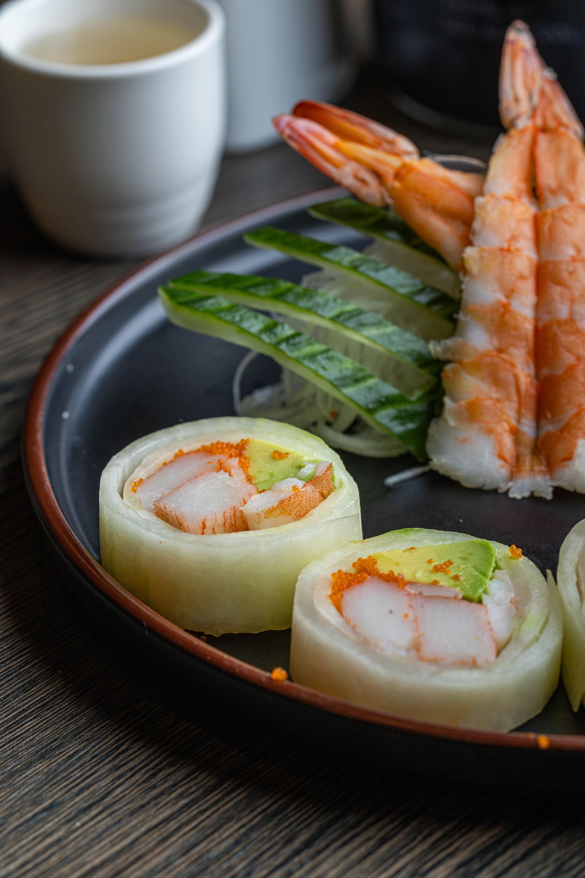 Sushi platter with shrimp, cucumber garnish, and two white fish rolls on a dark plate beside a cup
