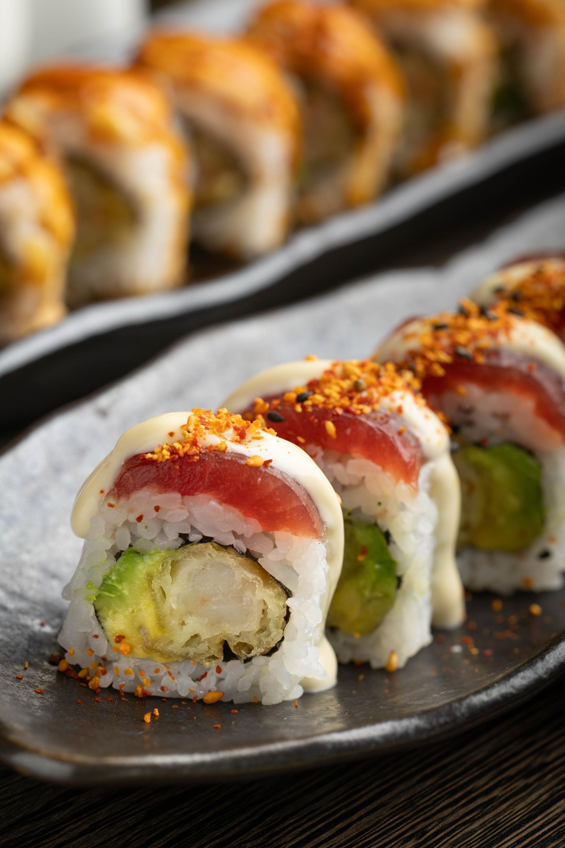 Sushi rolls topped with red sauce and seasoning on a black plate, with more rolls blurred in the background