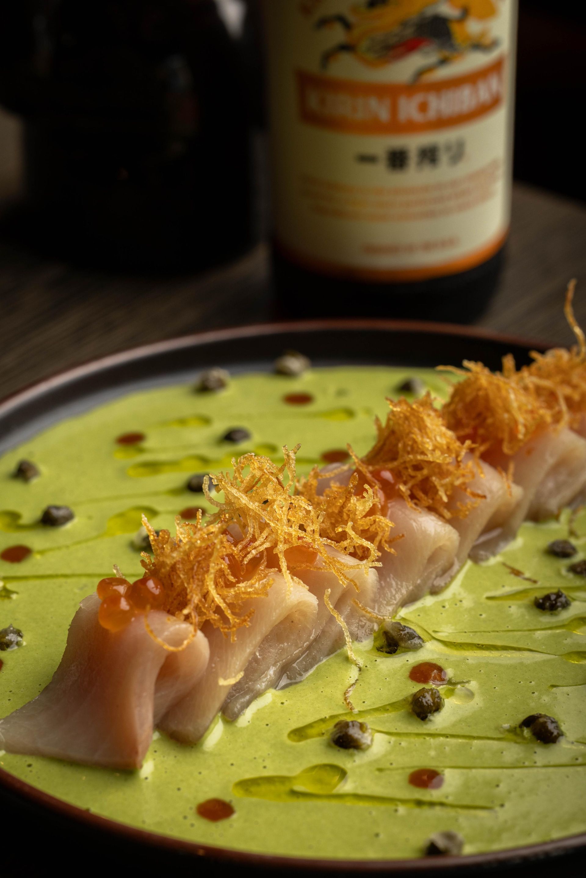 Sliced sashimi with green sauce and crispy garnish on a plate, with a bottle in the background