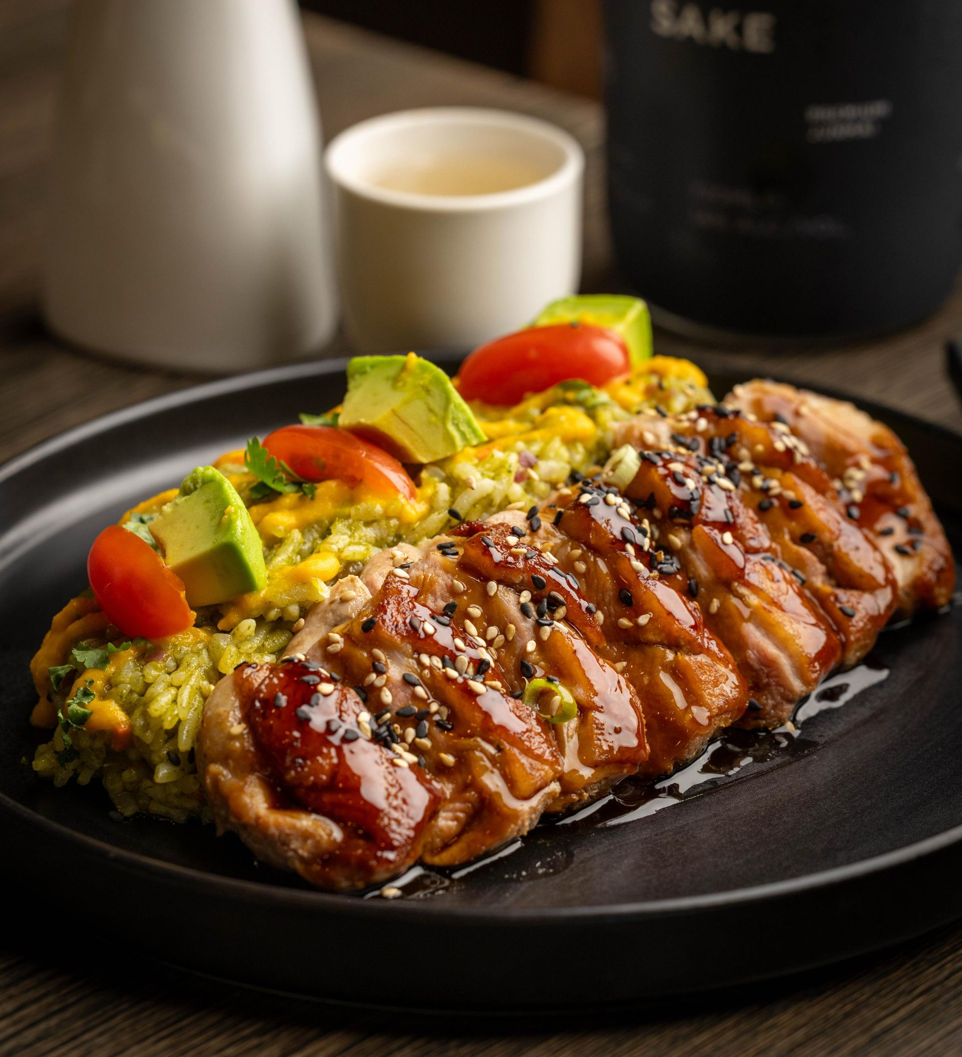 Glazed sliced meat on a black plate with avocado, tomatoes, and sauce, served on a table