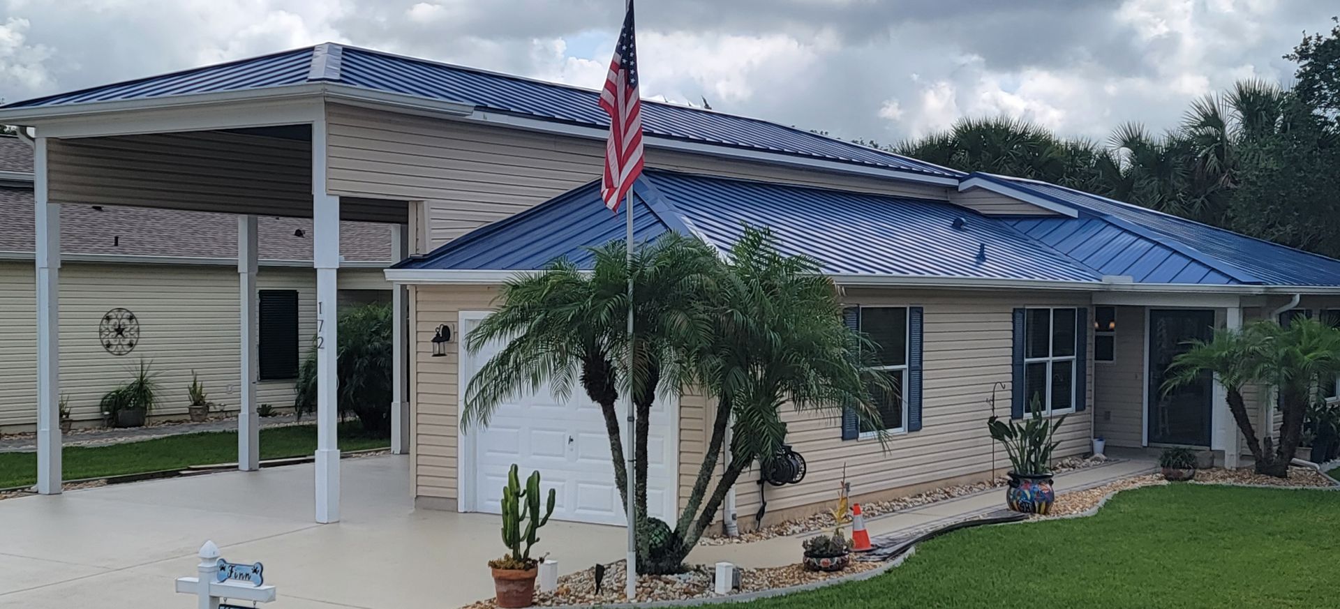 Single-story stone house with blue metal roof, covered porch, and an American flag by a palm tree.