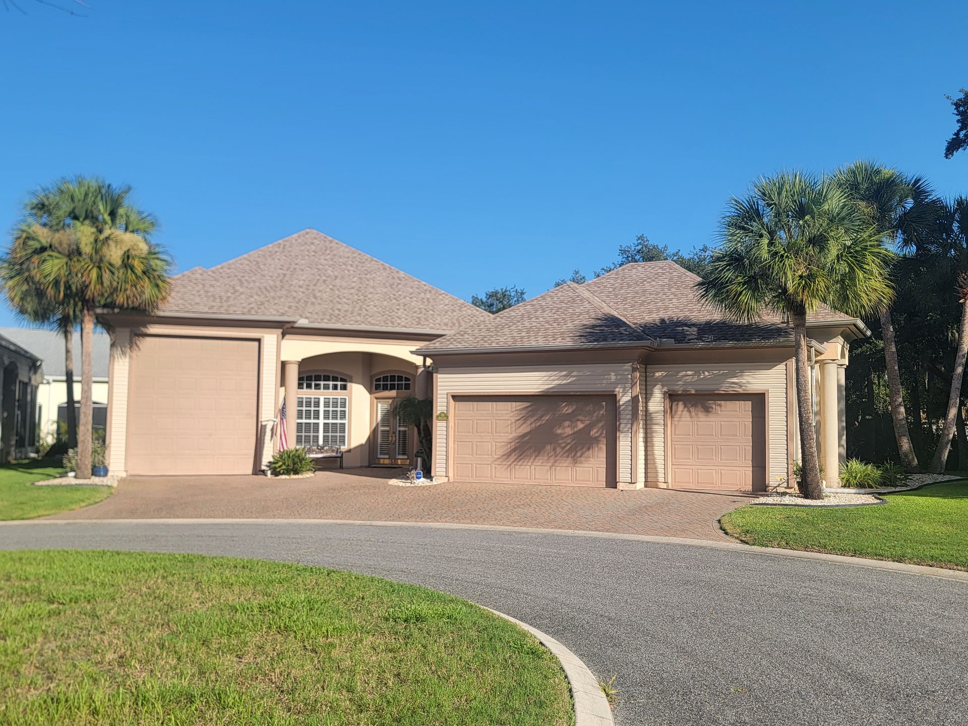 Single-story beige suburban house with a gray roof, driveway, and palm trees under a clear blue sky