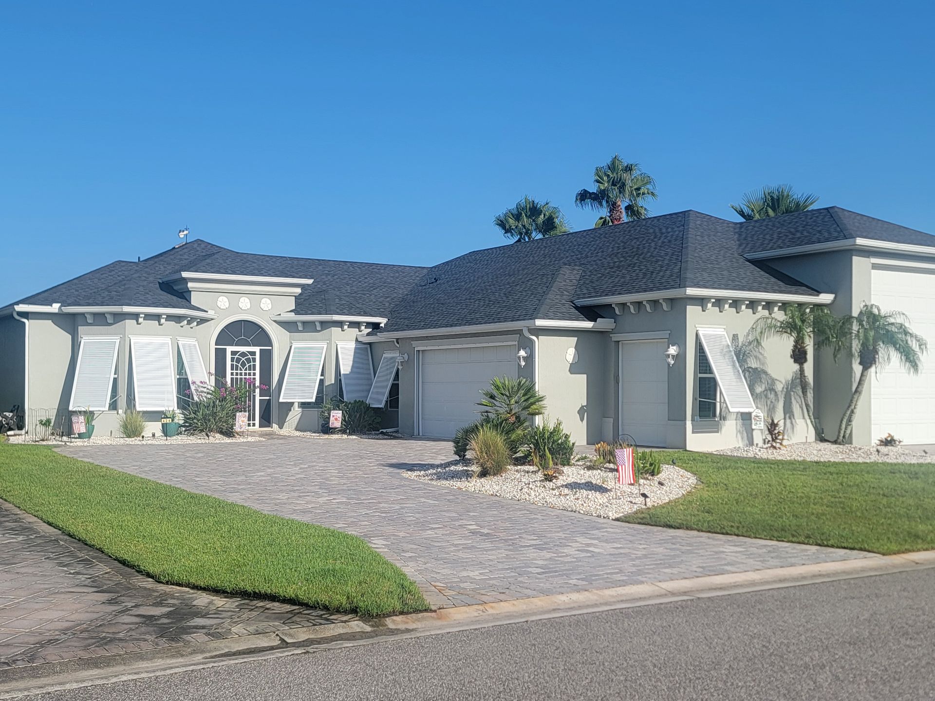 Single-story beige house with dark roof, stone driveway, and green lawn under a clear blue sky