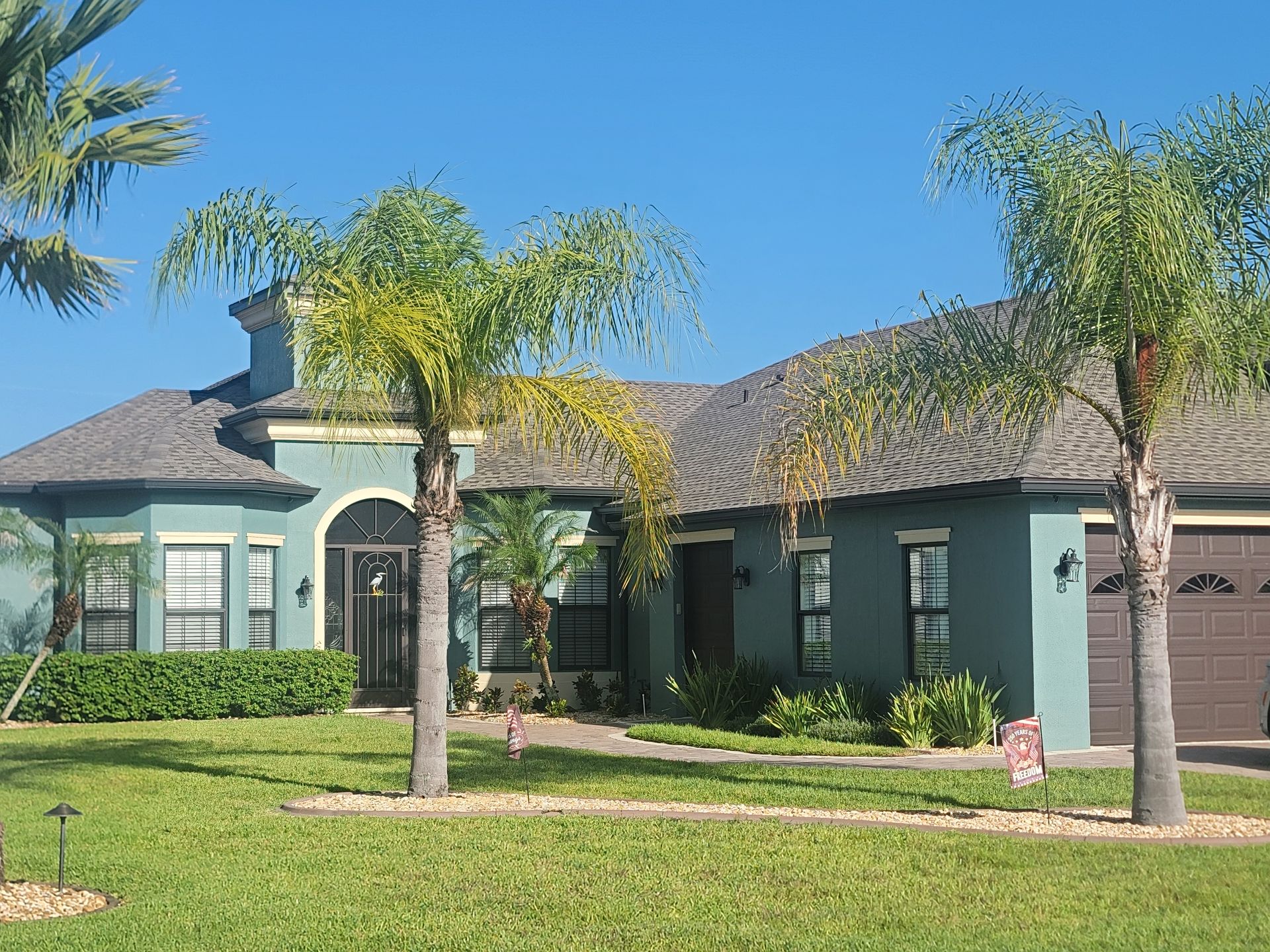 Single-story suburban house with palm trees, green lawn, and a two-car garage under a blue sky