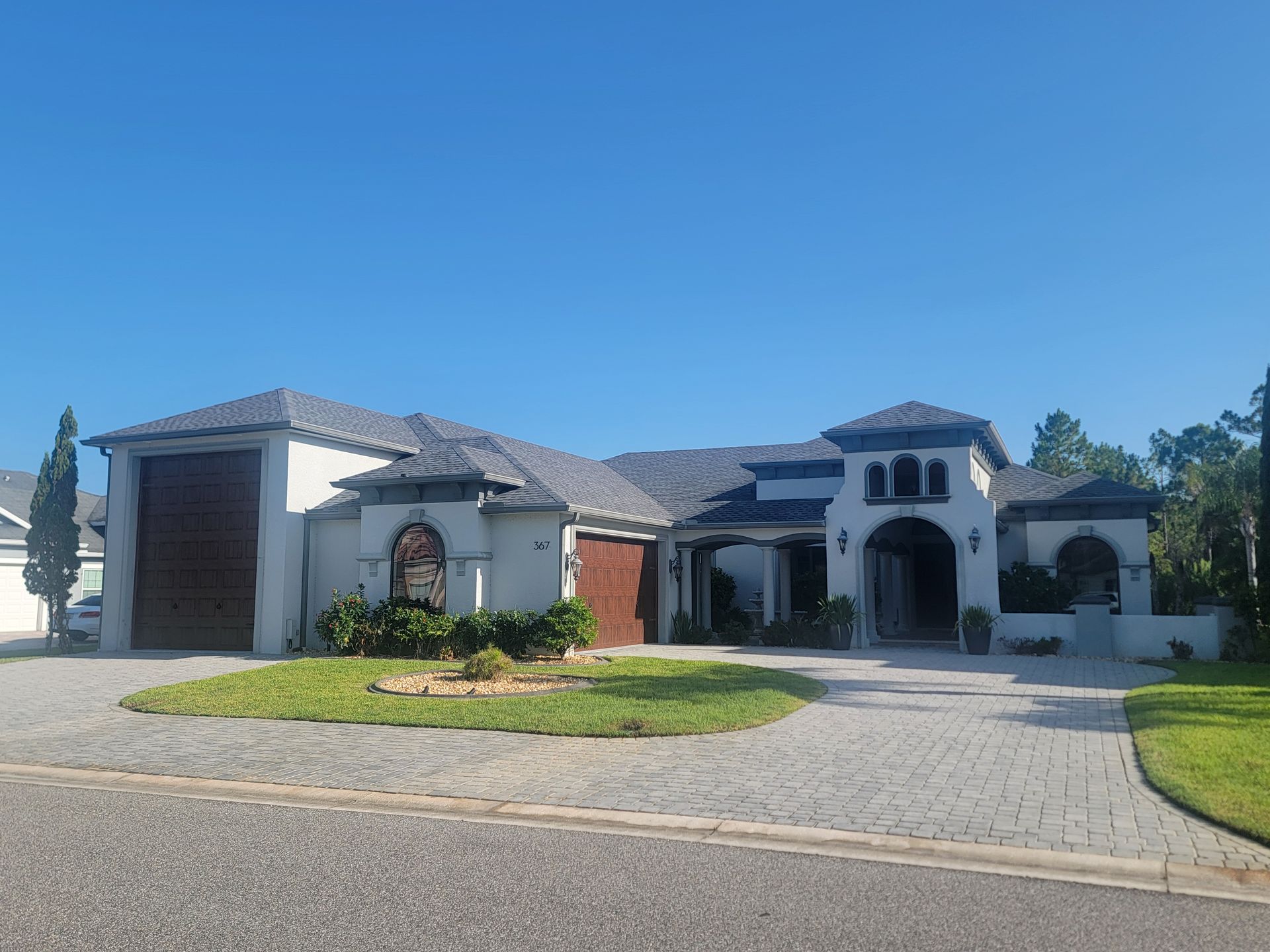 Large modern white house with a tiled roof, arched entry, and a paved driveway under a clear blue sky