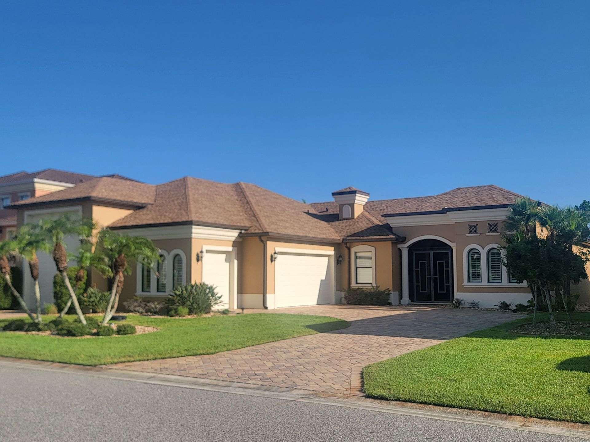 Large beige stucco house with a tiled roof, garage, and green lawn under a clear blue sky