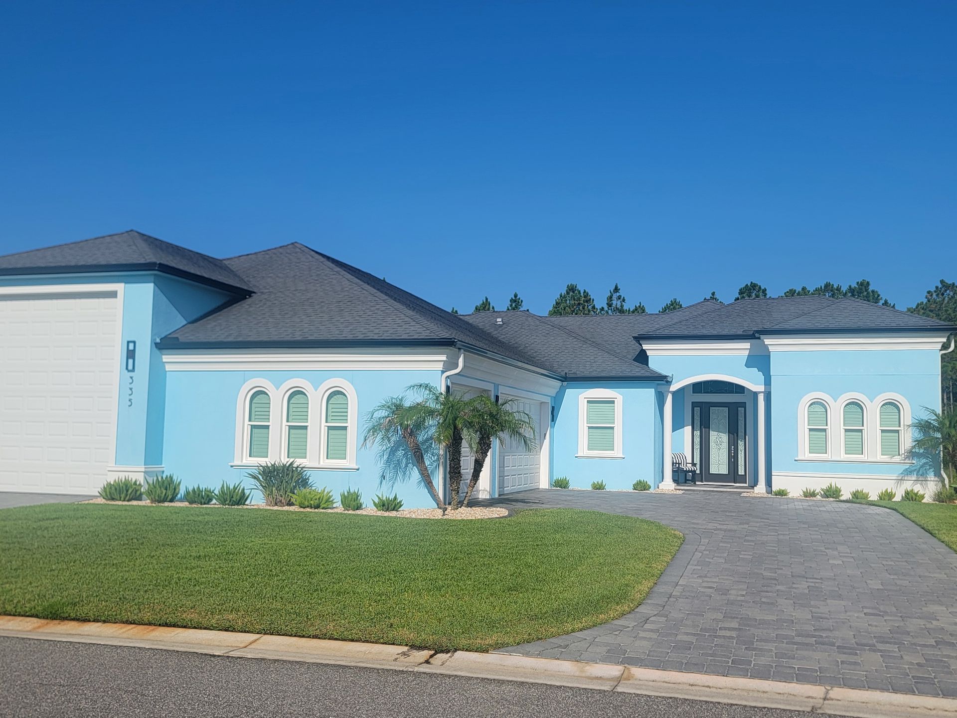 Blue single-story house with arched windows, gray roof, green lawn, and curved driveway under a clear sky