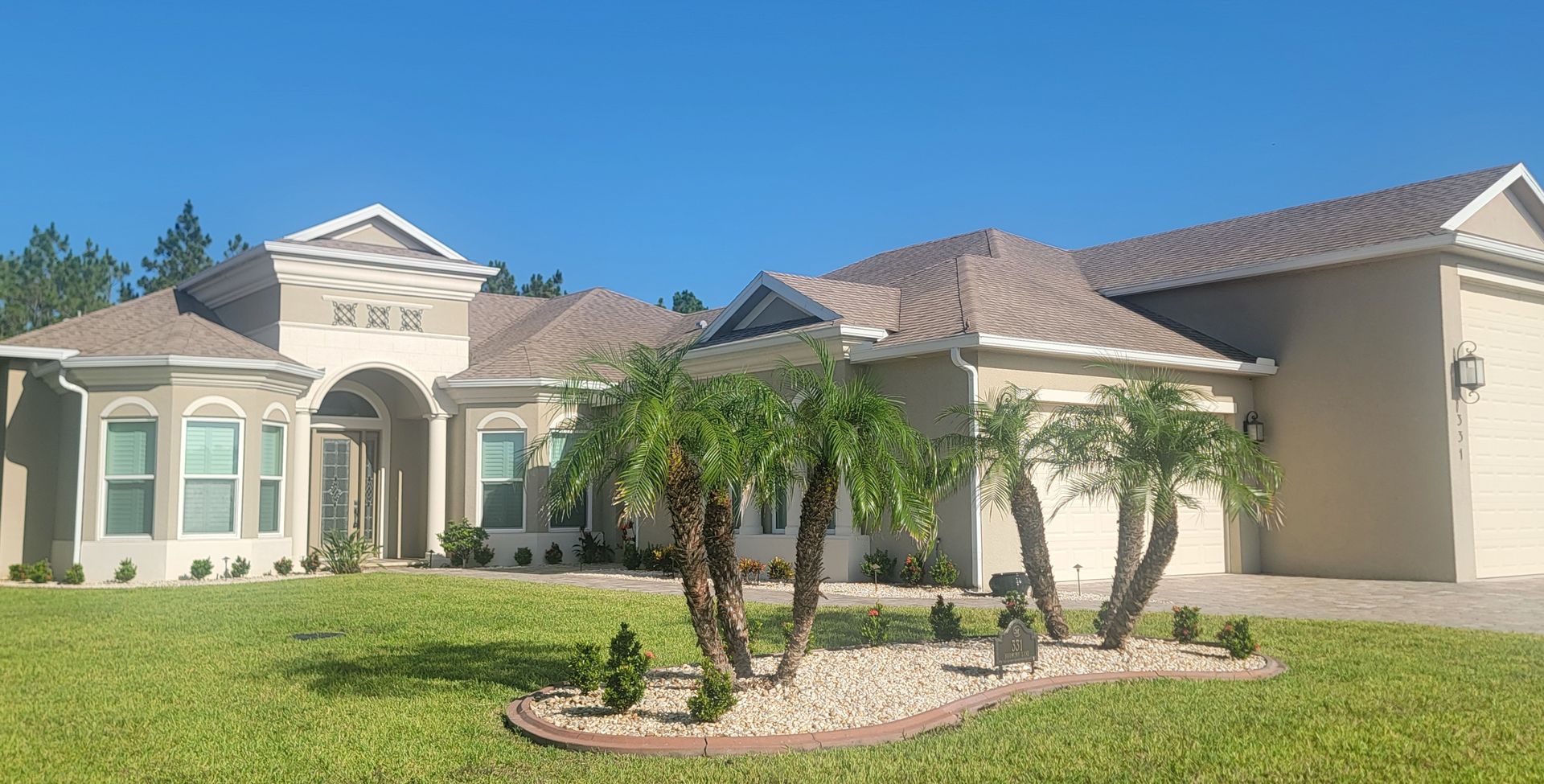 Single-story suburban house with a tile roof, palm trees, and a green lawn in bright sunlight
