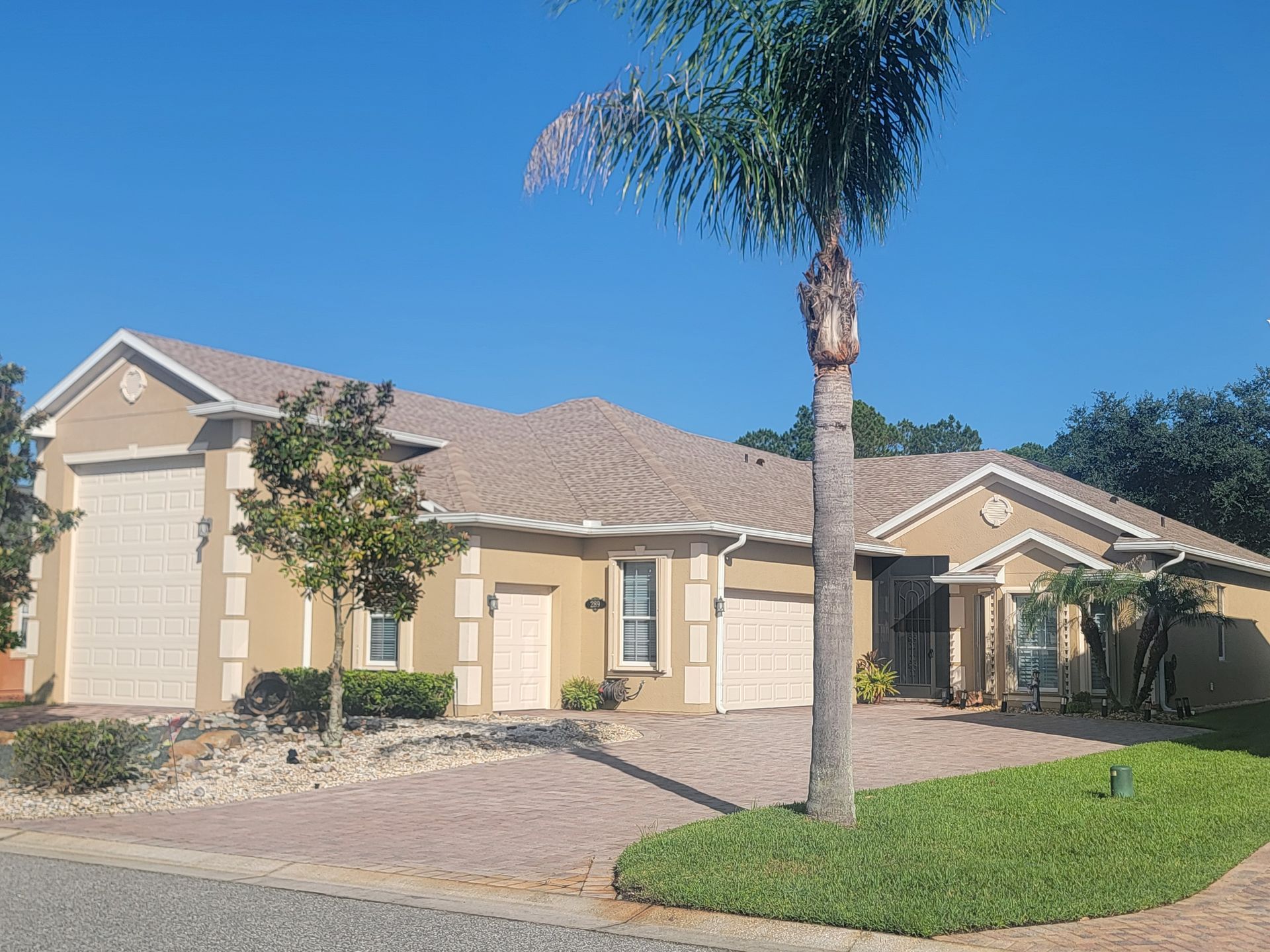 Single-story beige suburban house with tiled roof, palm tree, and paved driveway under a blue sky