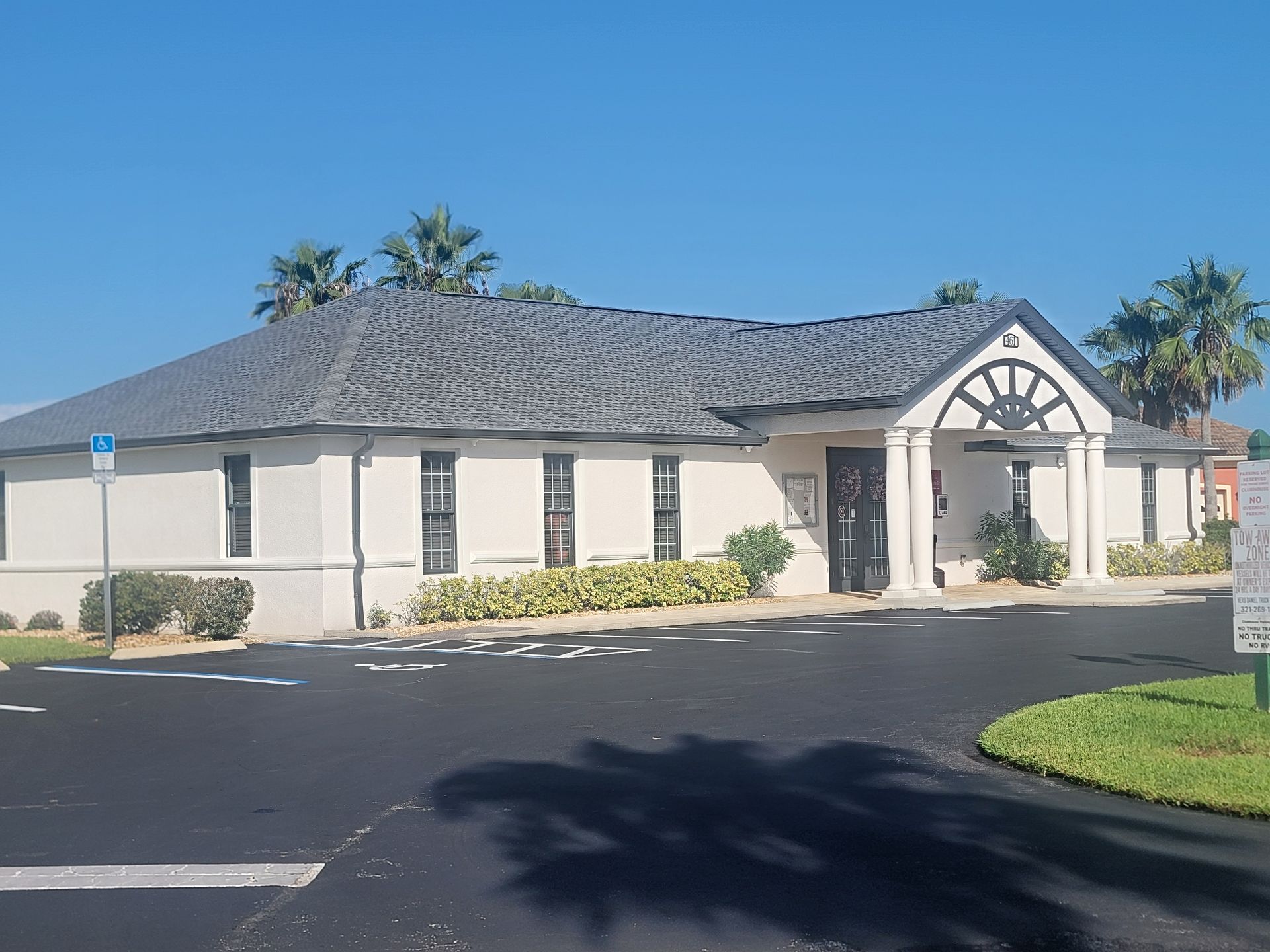 Single-story beige building with gray roof, arched entrance, and palm trees on a sunny day