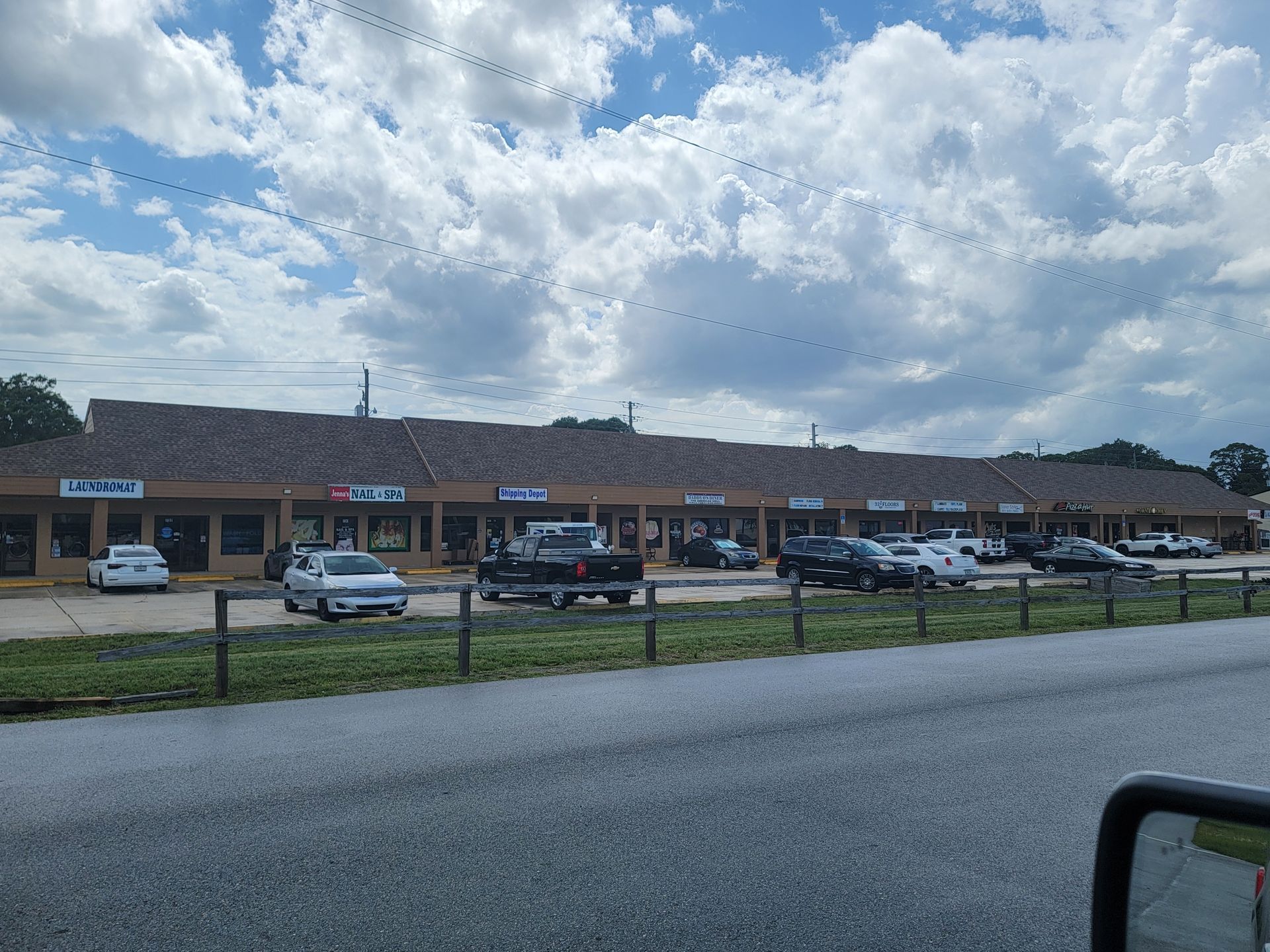 Strip mall storefronts with parked cars under a cloudy sky, viewed from across a road.