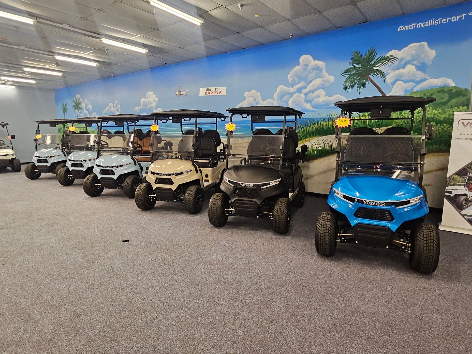 Row of colorful golf carts displayed indoors in a showroom with a mural sky backdrop