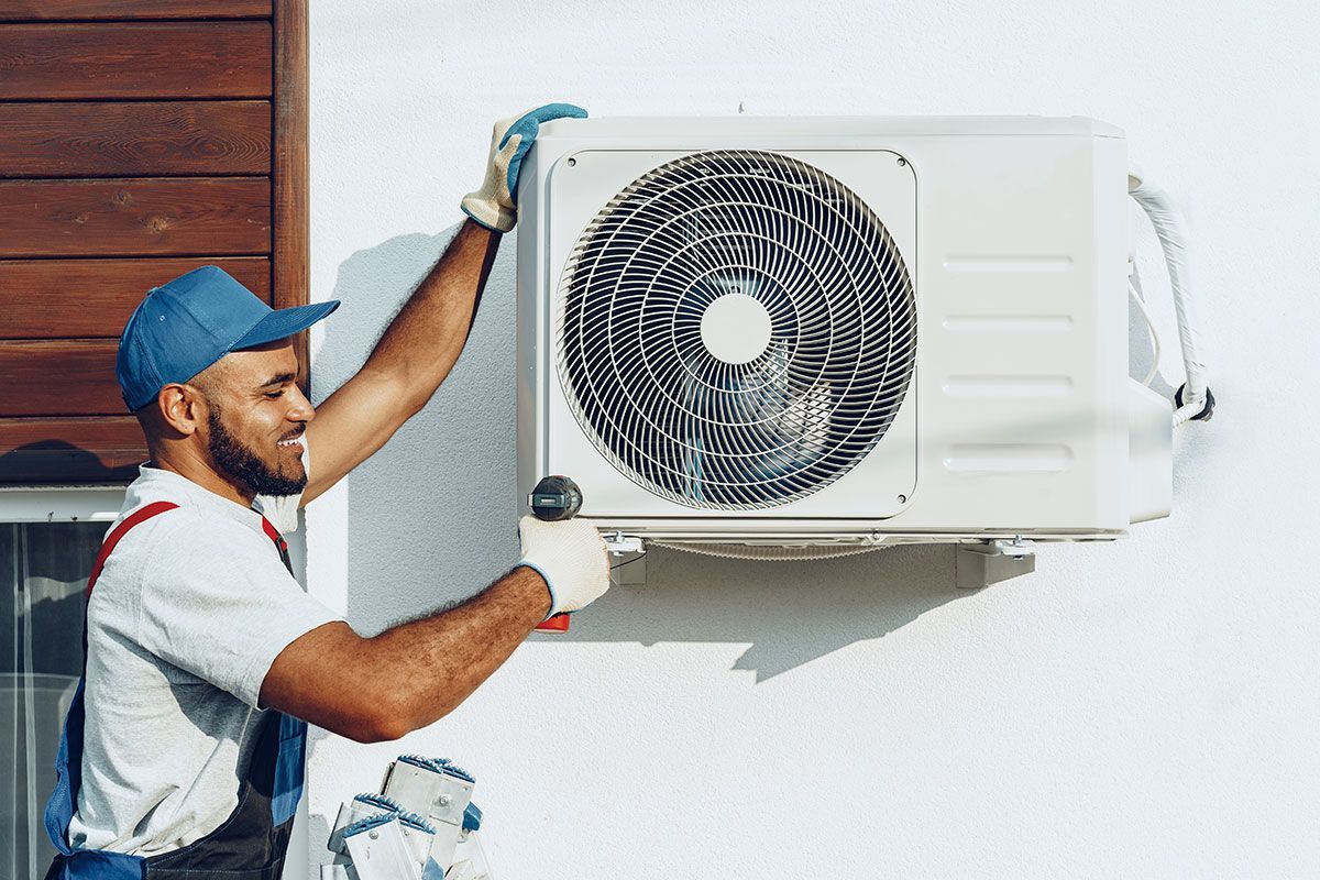 Person wearing a blue hat and gloves installing an AC unit on a white wall.